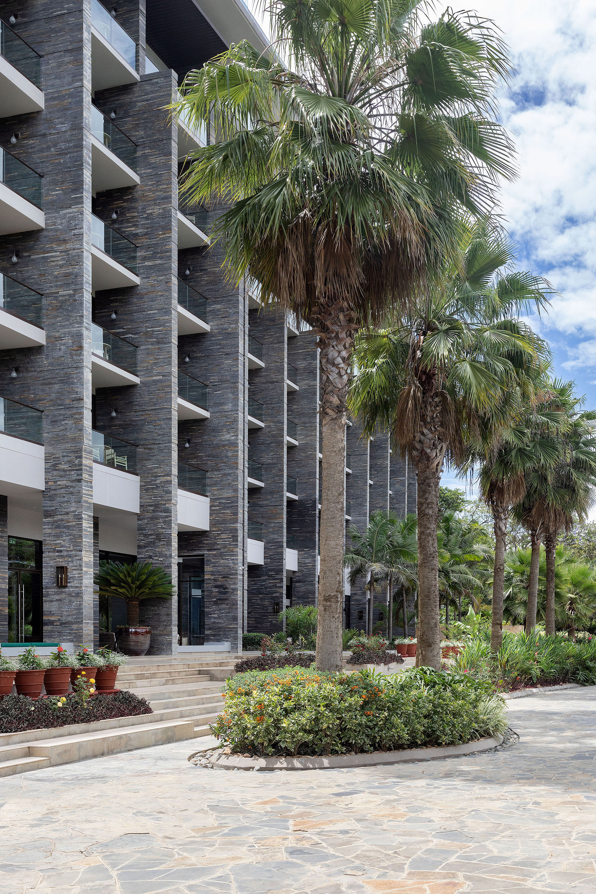 Modern resort building, stone facade, balconies, lush palm trees, patio.