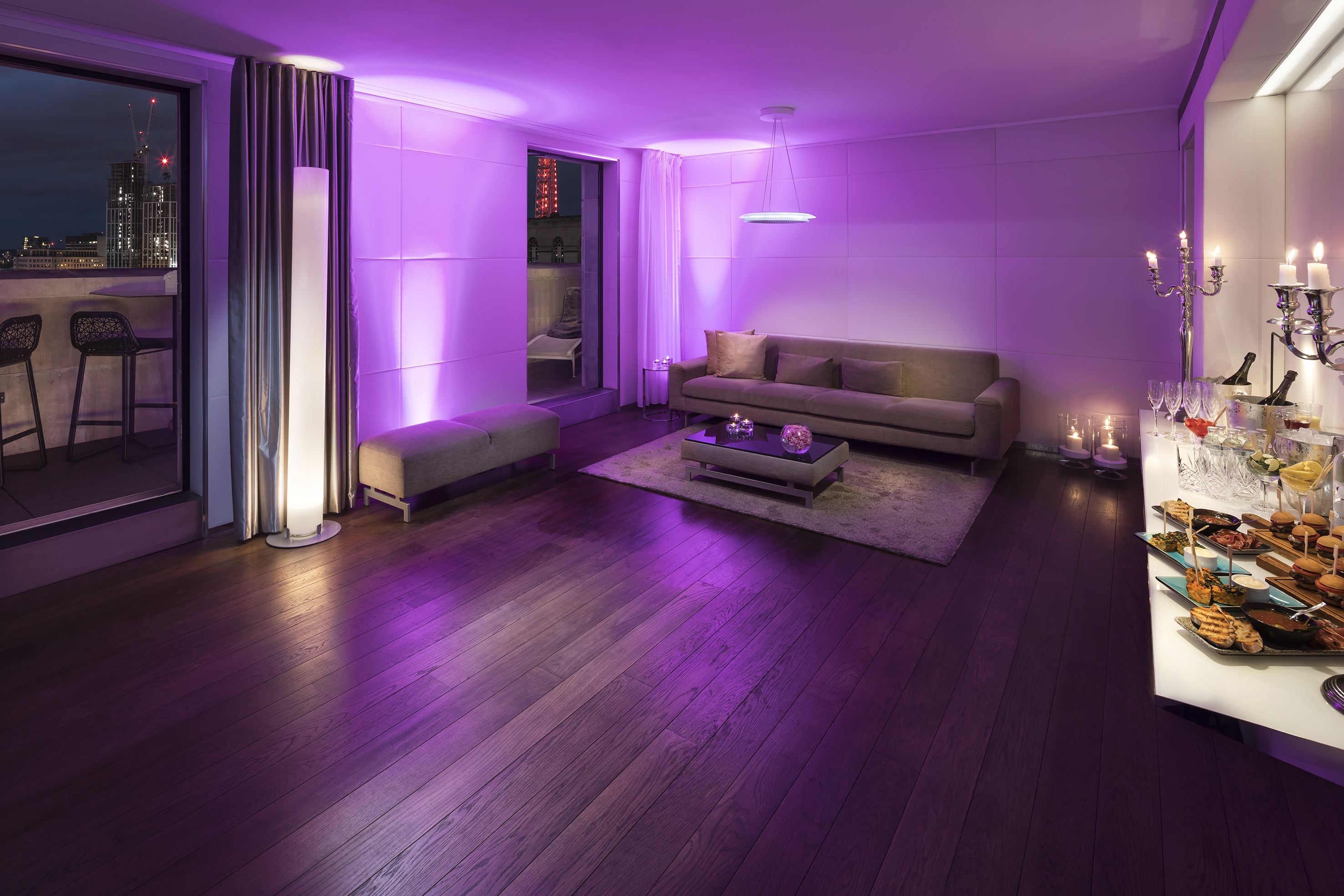 Modern lounge with purple ambient lighting, city view balcony, and buffet table.