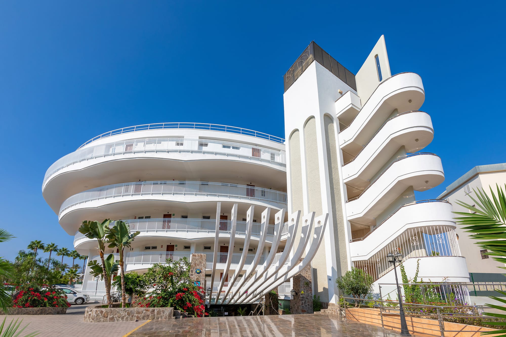 Modern white hotel with curved balconies, unique facade, and palm trees.