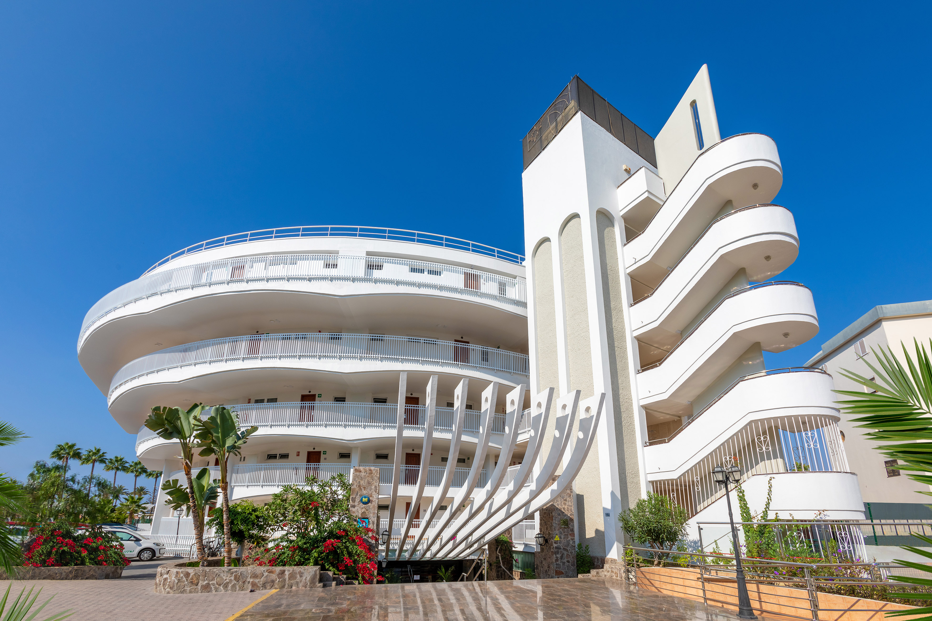 Modern white hotel with curved balconies, unique facade, and palm trees.