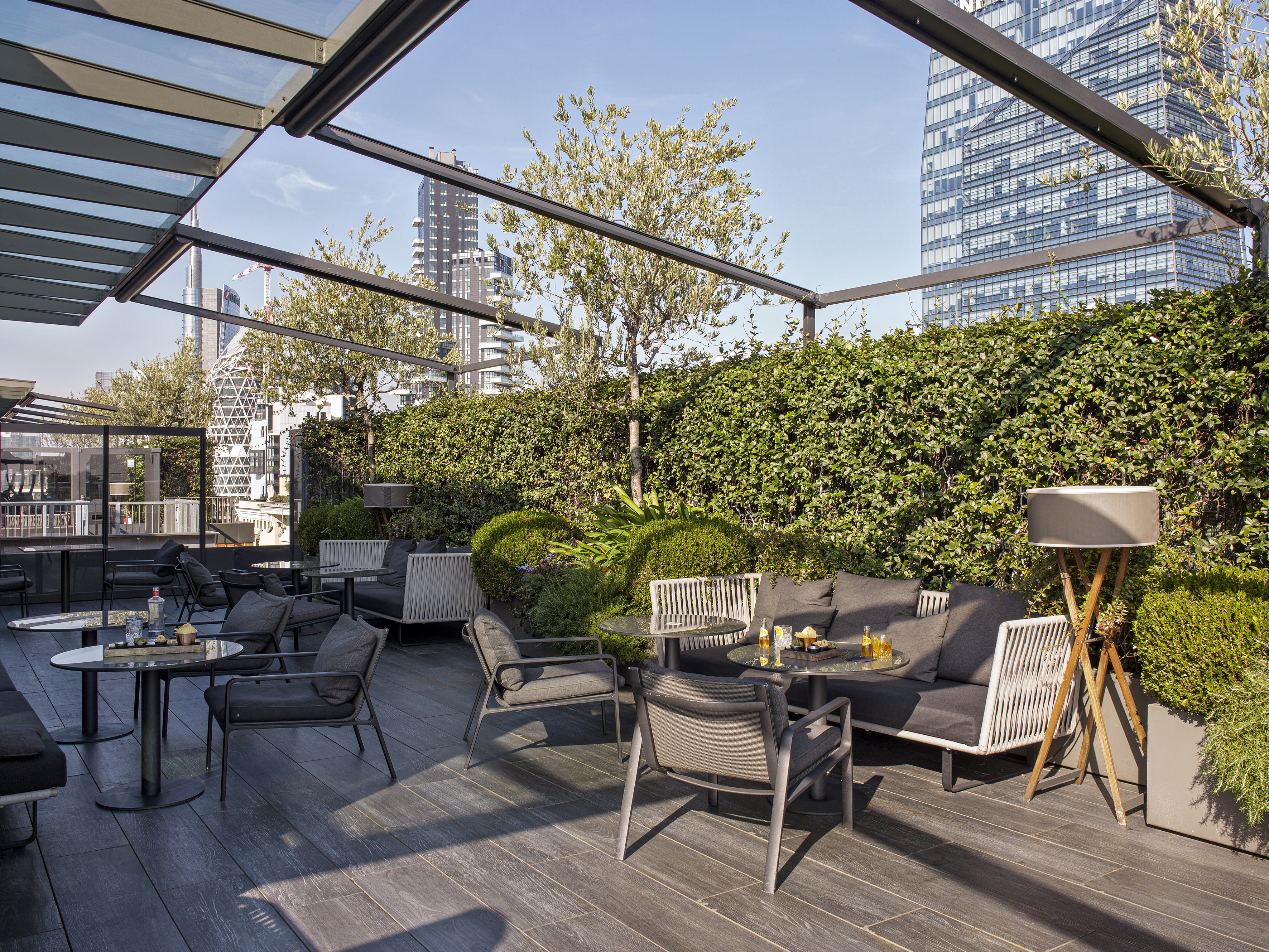 Modern outdoor lounge terrace with grey seating, green hedges, and city skyline.