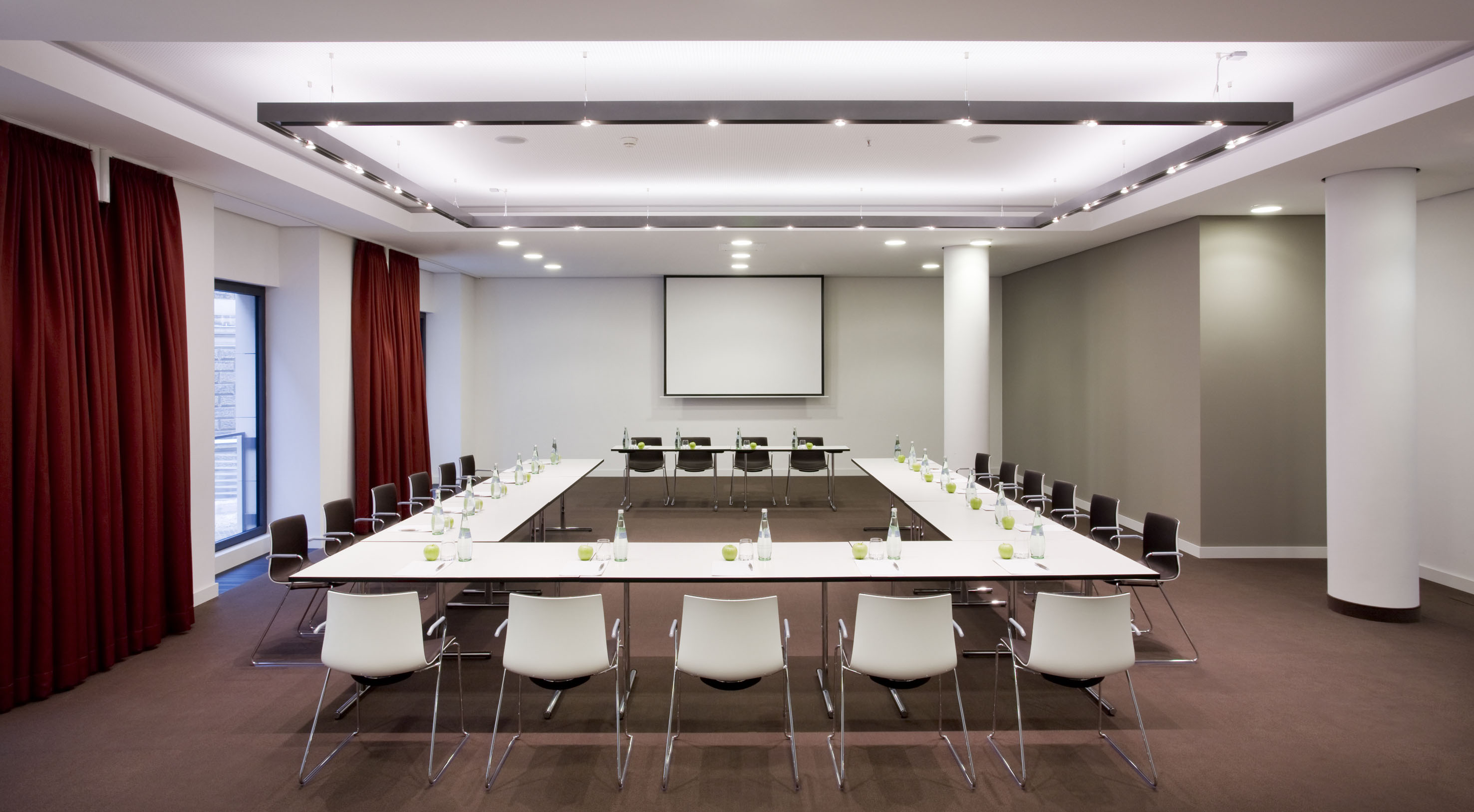 Modern conference room with U-shaped table, projection screen, and red curtains.