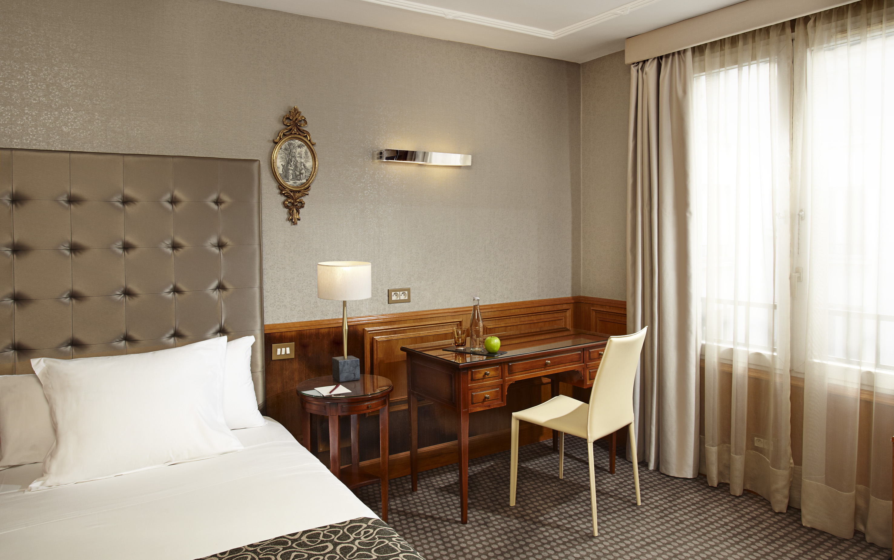 Elegant hotel room: upholstered bed, wooden desk, lamp, and window.