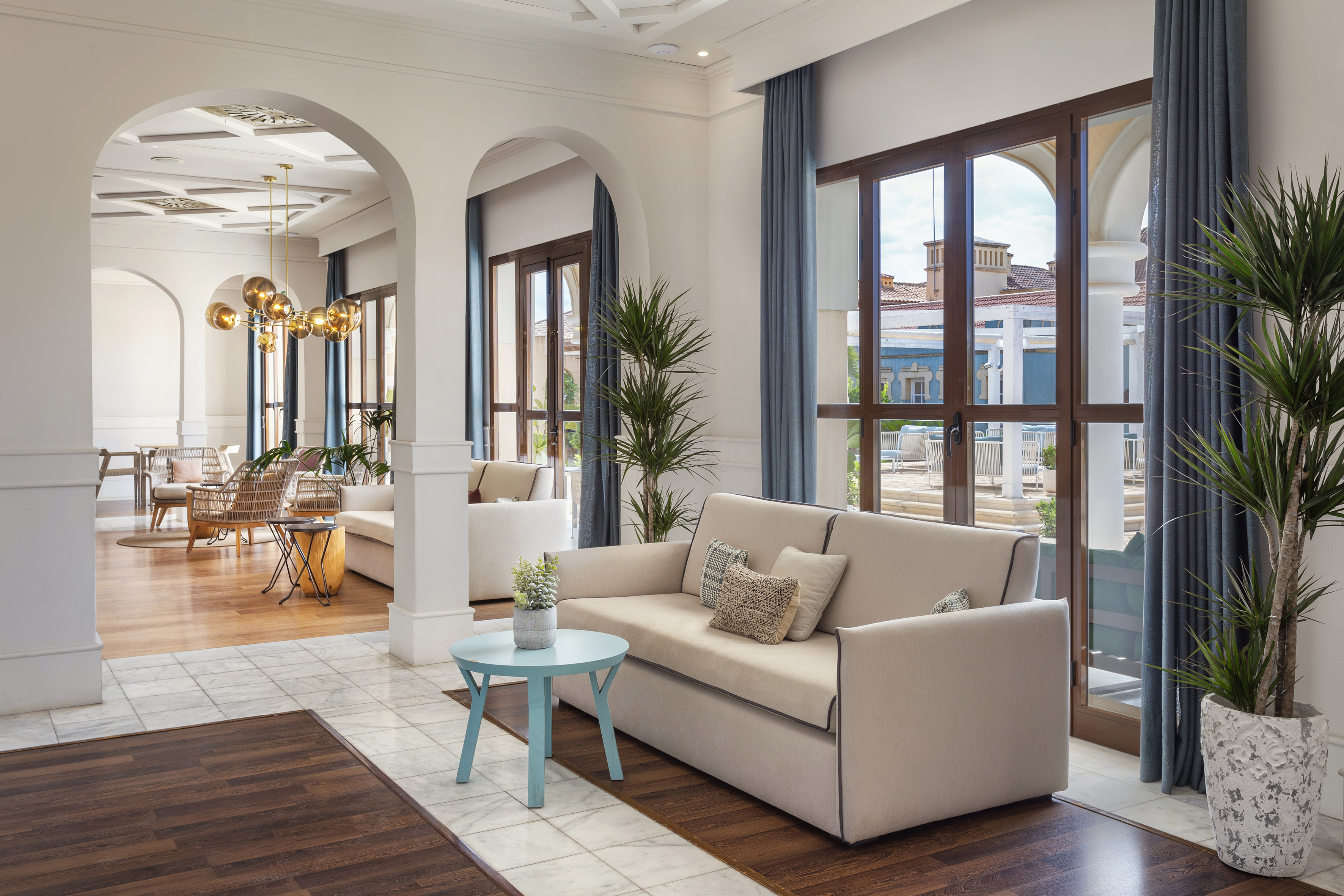 Modern hotel lobby with beige sofa, blue table, large windows, and arched entry.