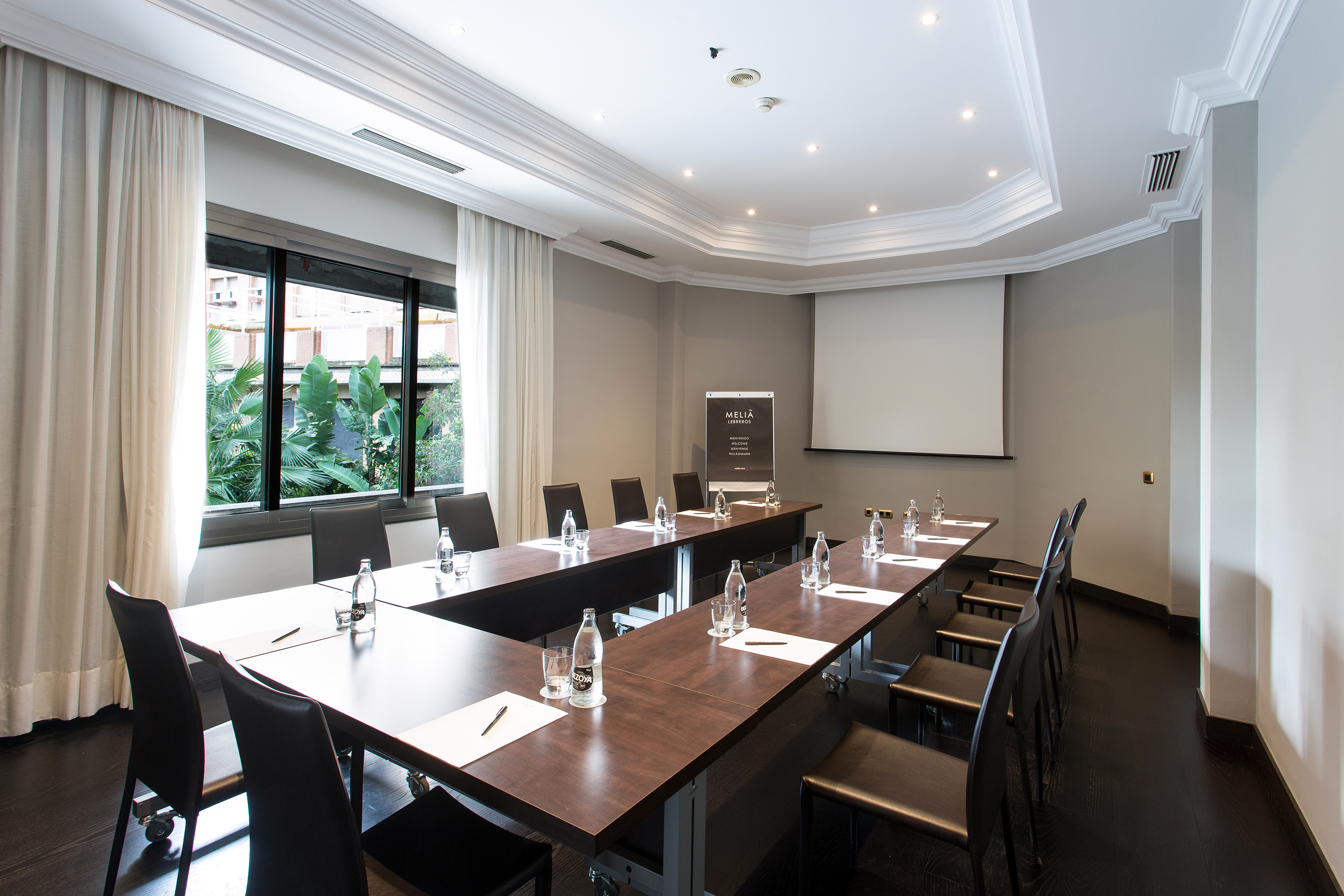 Modern Meliá hotel meeting room with U-table setup, projector screen, and window.