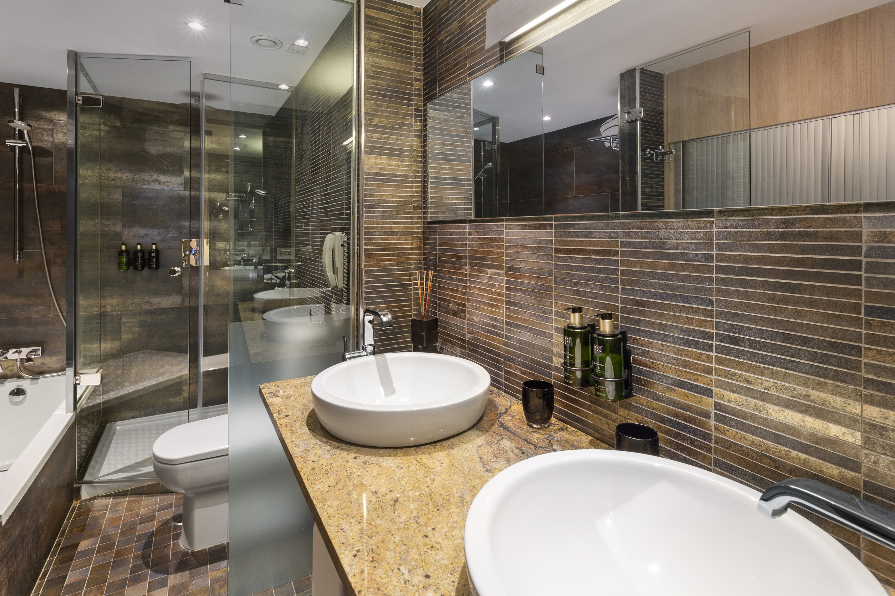 Hotel bathroom with two vessel sinks, granite counter, and textured brown tiles.