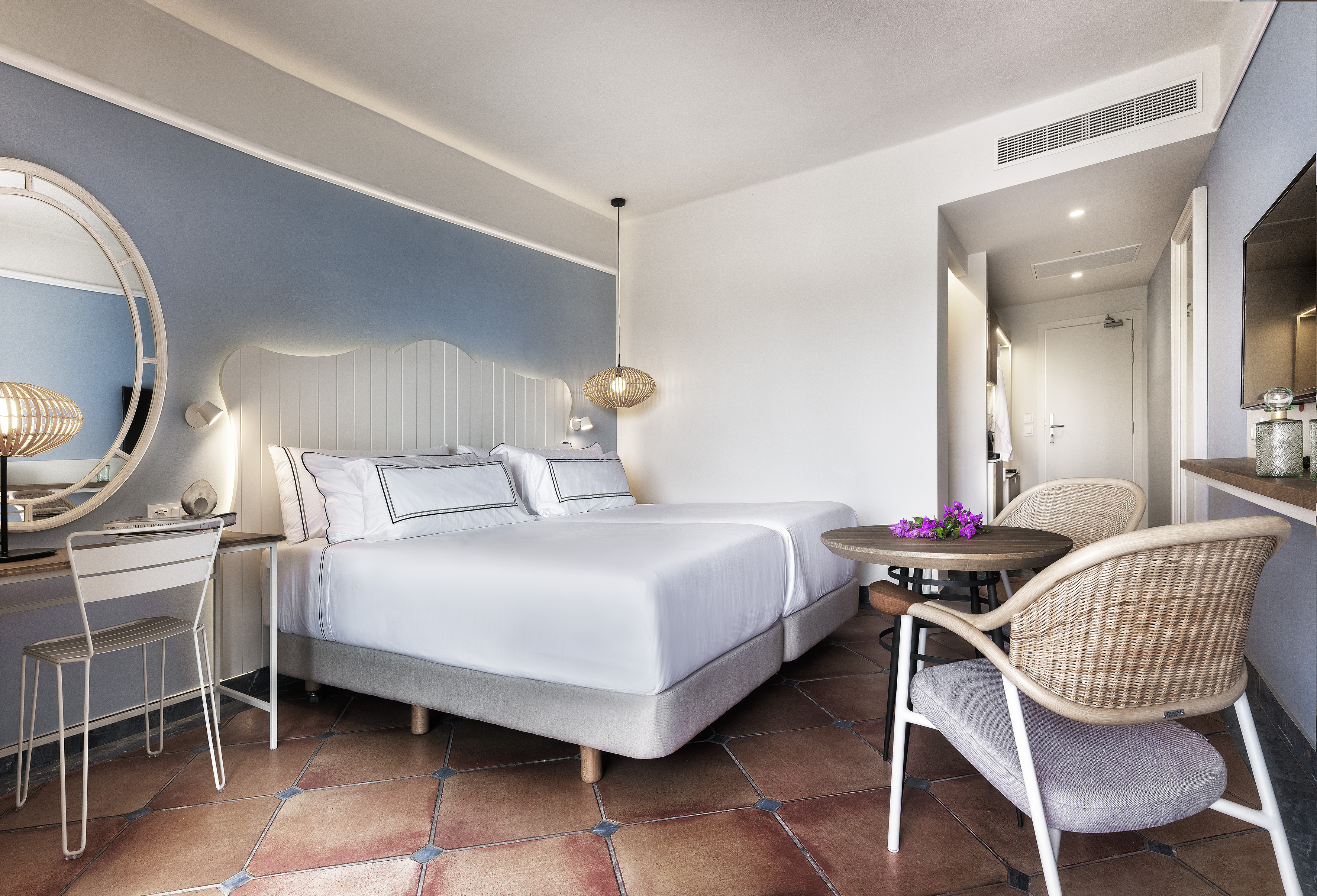 Modern hotel bedroom with twin beds, blue accent wall, and rattan furniture.