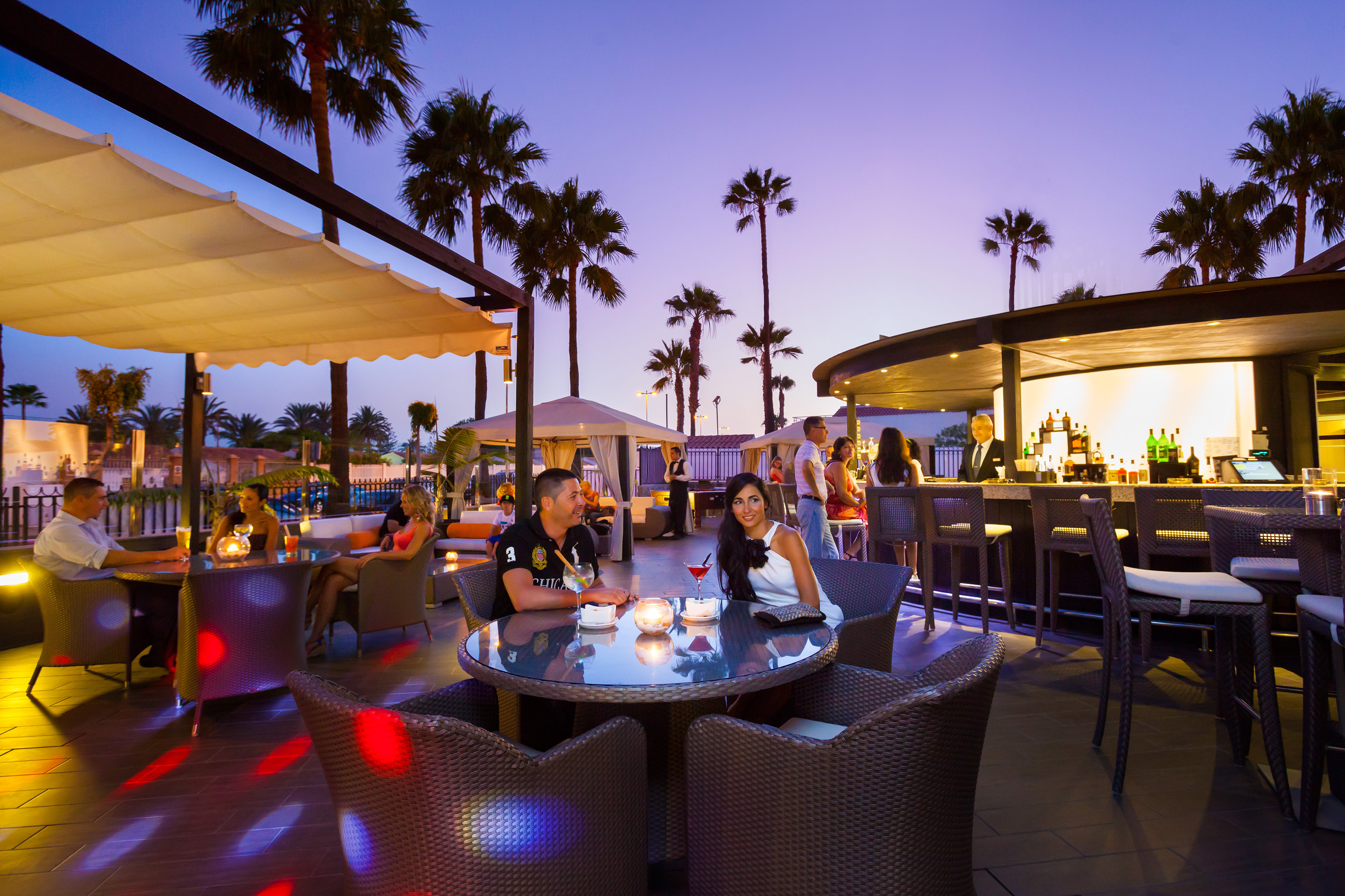 Outdoor resort bar with guests enjoying drinks at dusk, surrounded by palm trees.