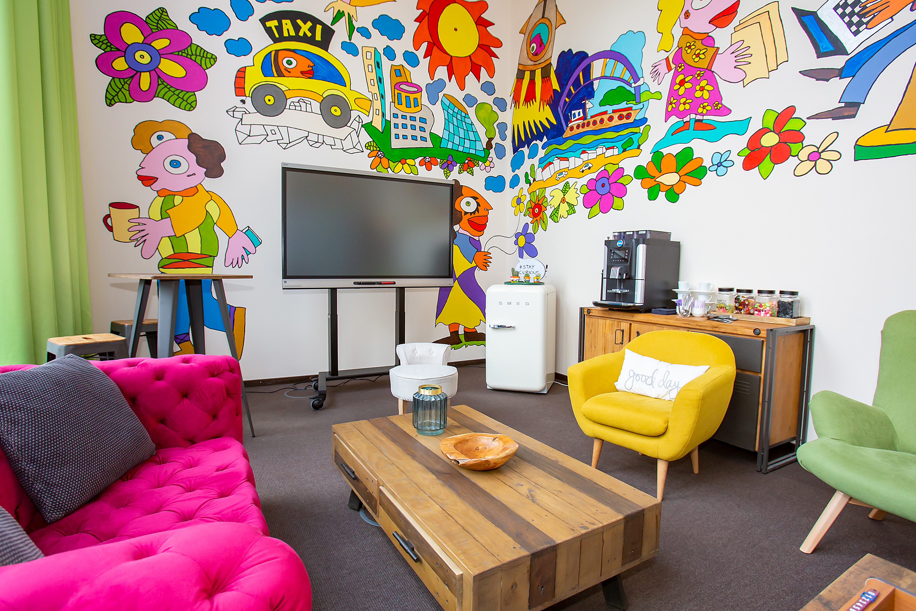 Colorful lounge with playful cartoon wall mural, pink sofa, coffee station, and TV.
