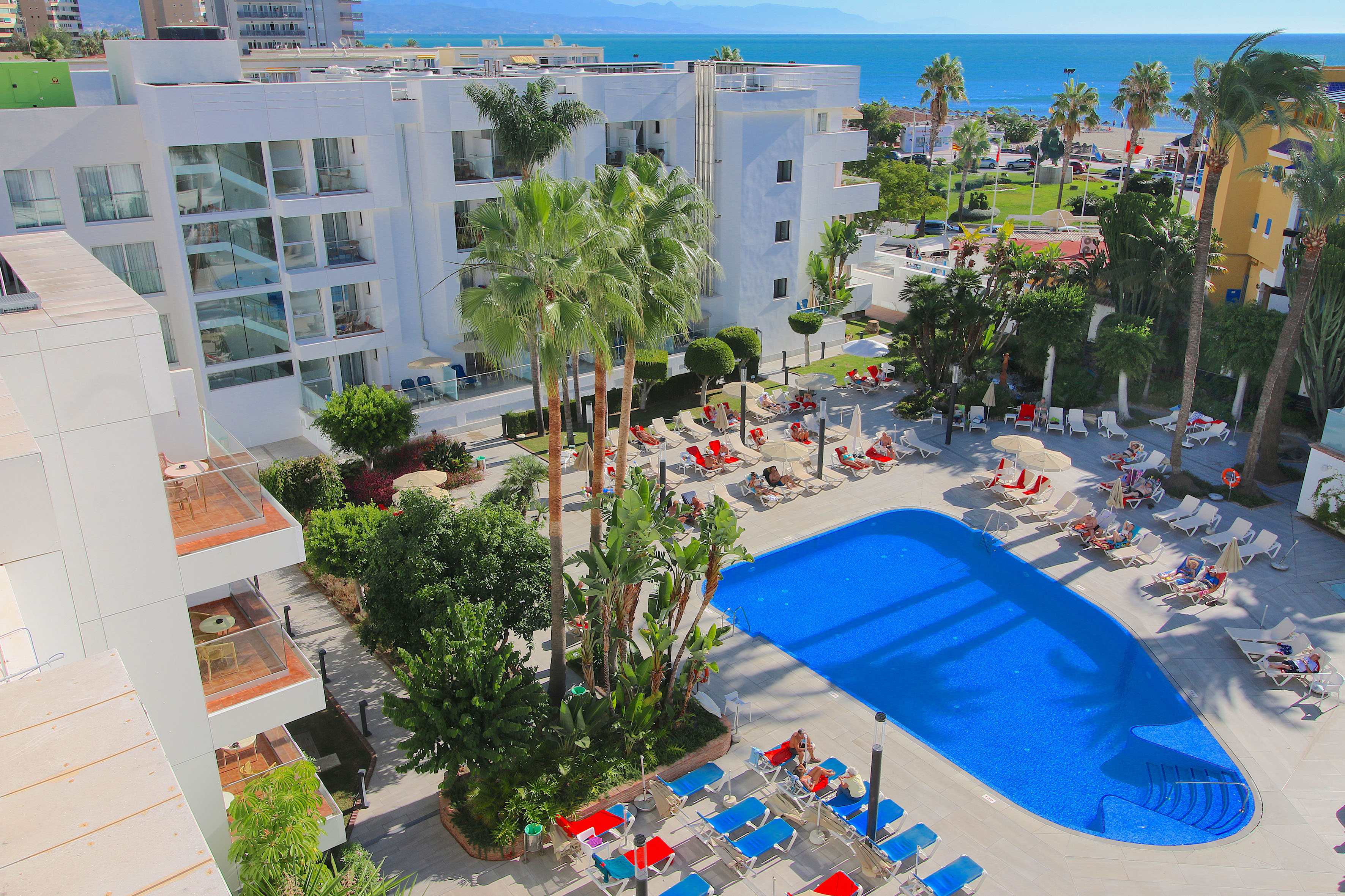 Aerial view of sunny hotel resort, blue pool, palm trees, beach, and ocean.