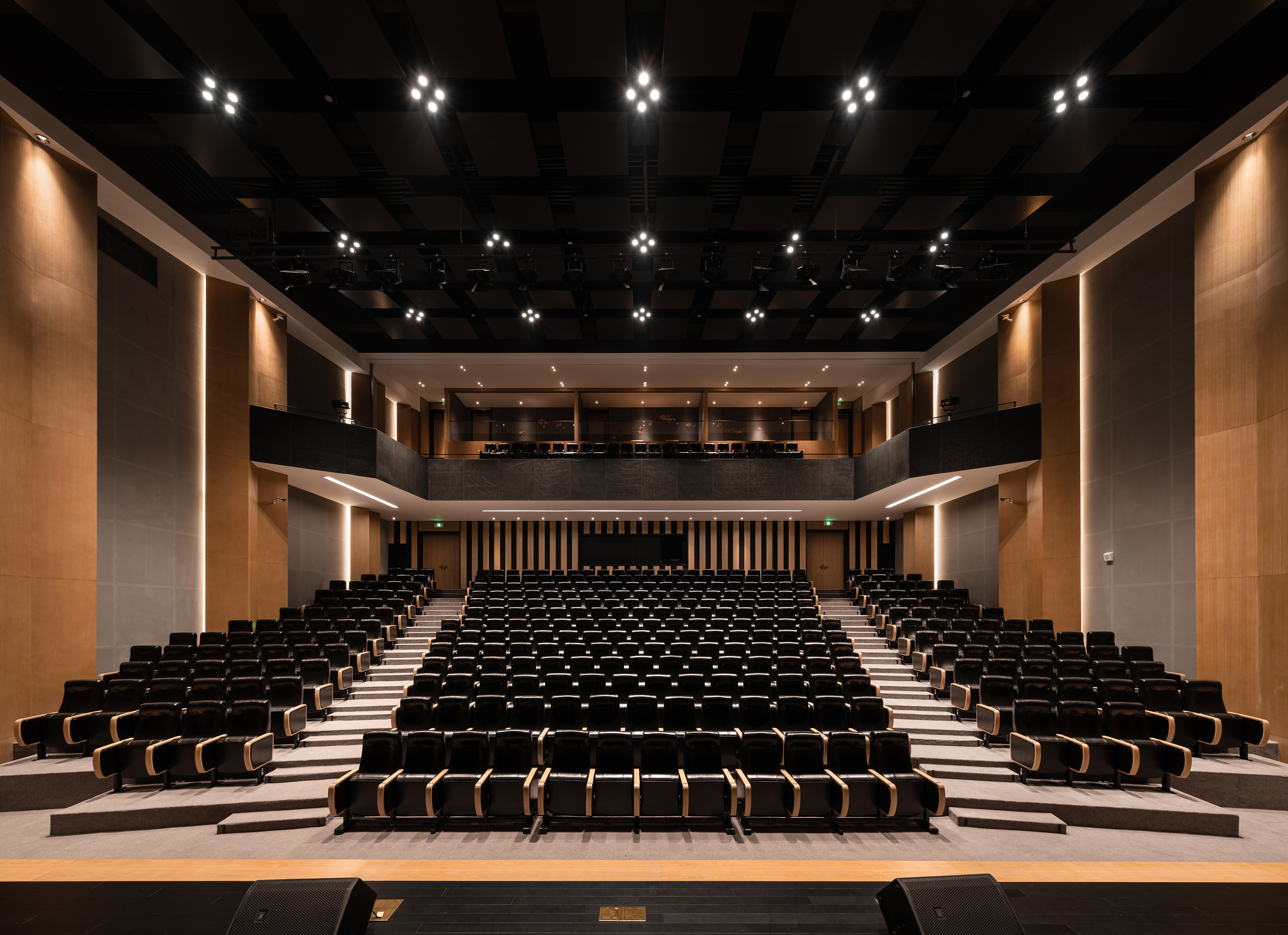 Empty modern auditorium with tiered black seating, wood acoustic panels, and stage.