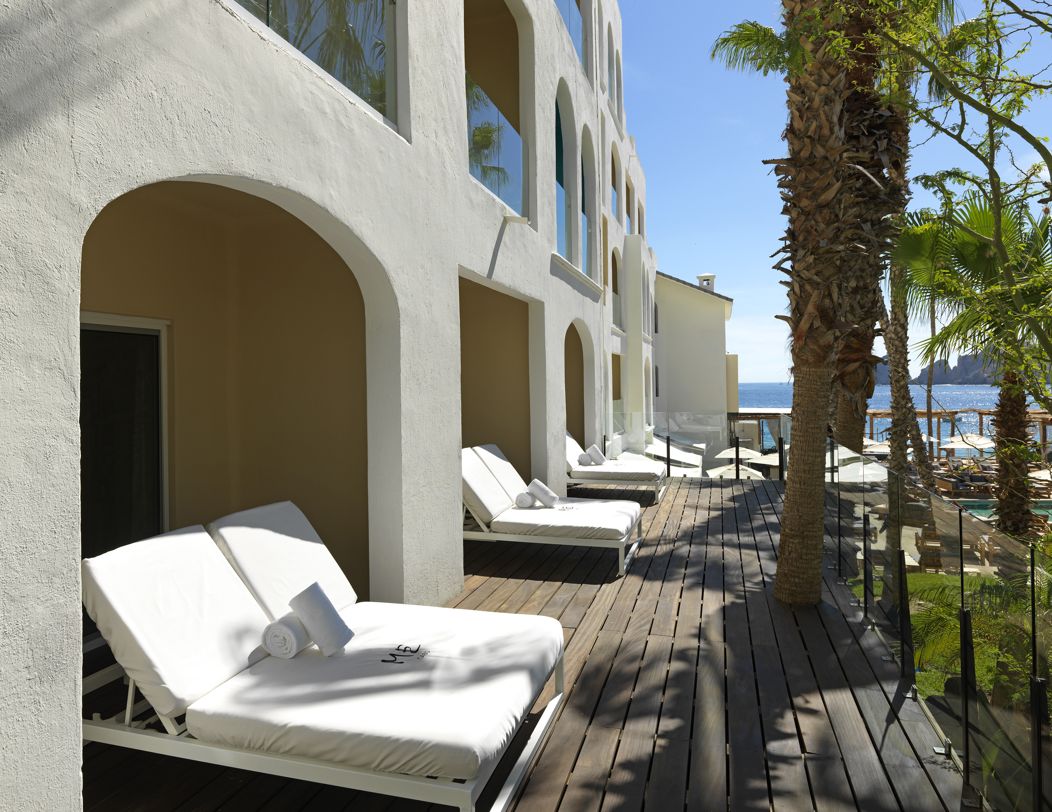Hotel balcony with white daybeds, archways, palm trees, and ocean view.