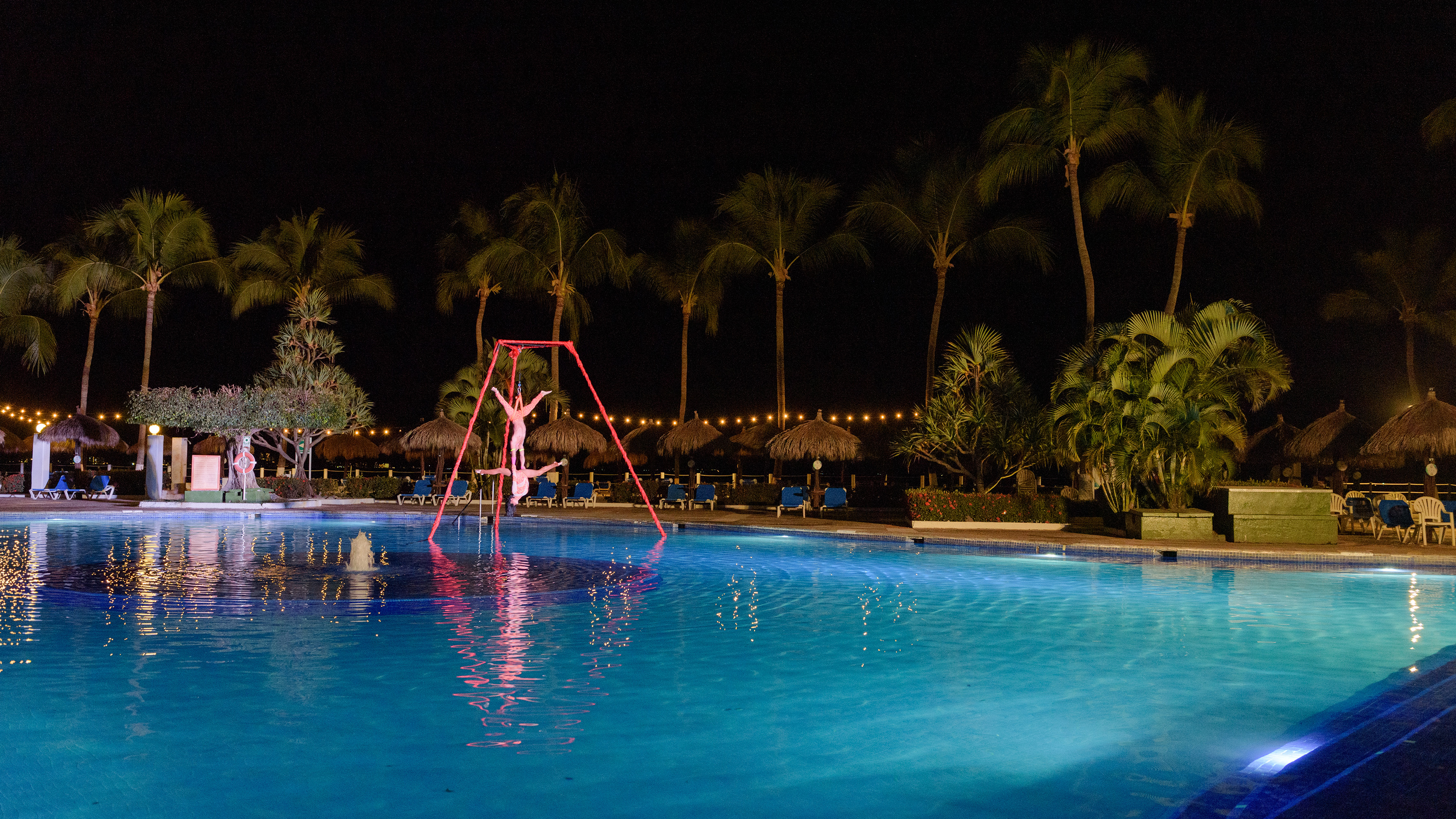 Night aerial acrobatics show over illuminated resort pool and palm trees.