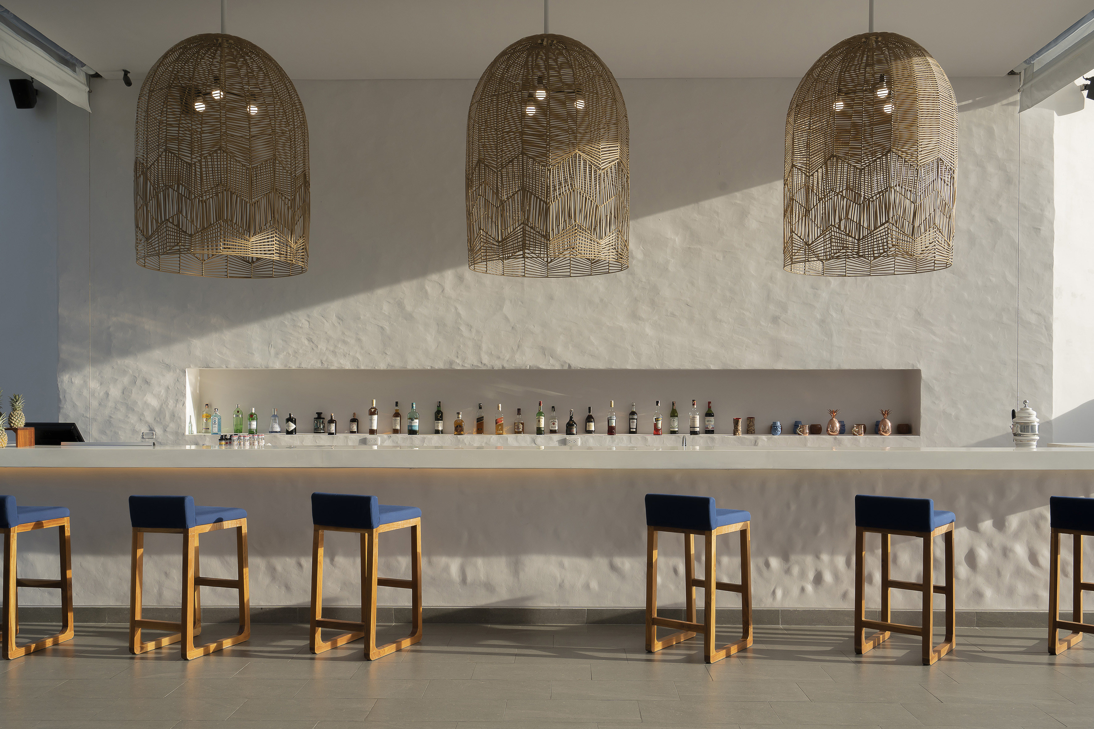 Stylish hotel bar with rattan pendant lights, blue stools, and liquor bottles.