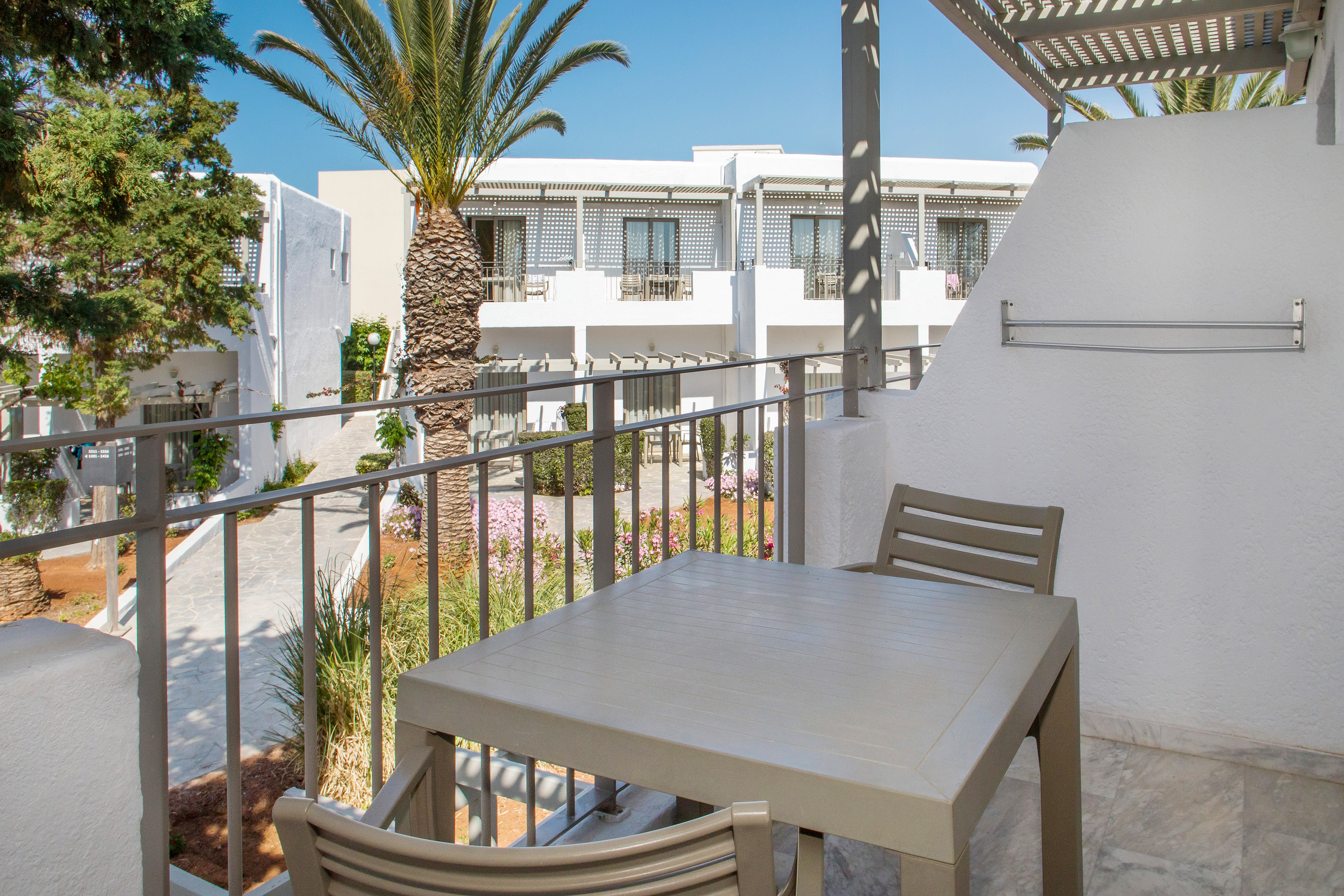 Hotel balcony with table and chairs, overlooking white resort buildings and palm trees.