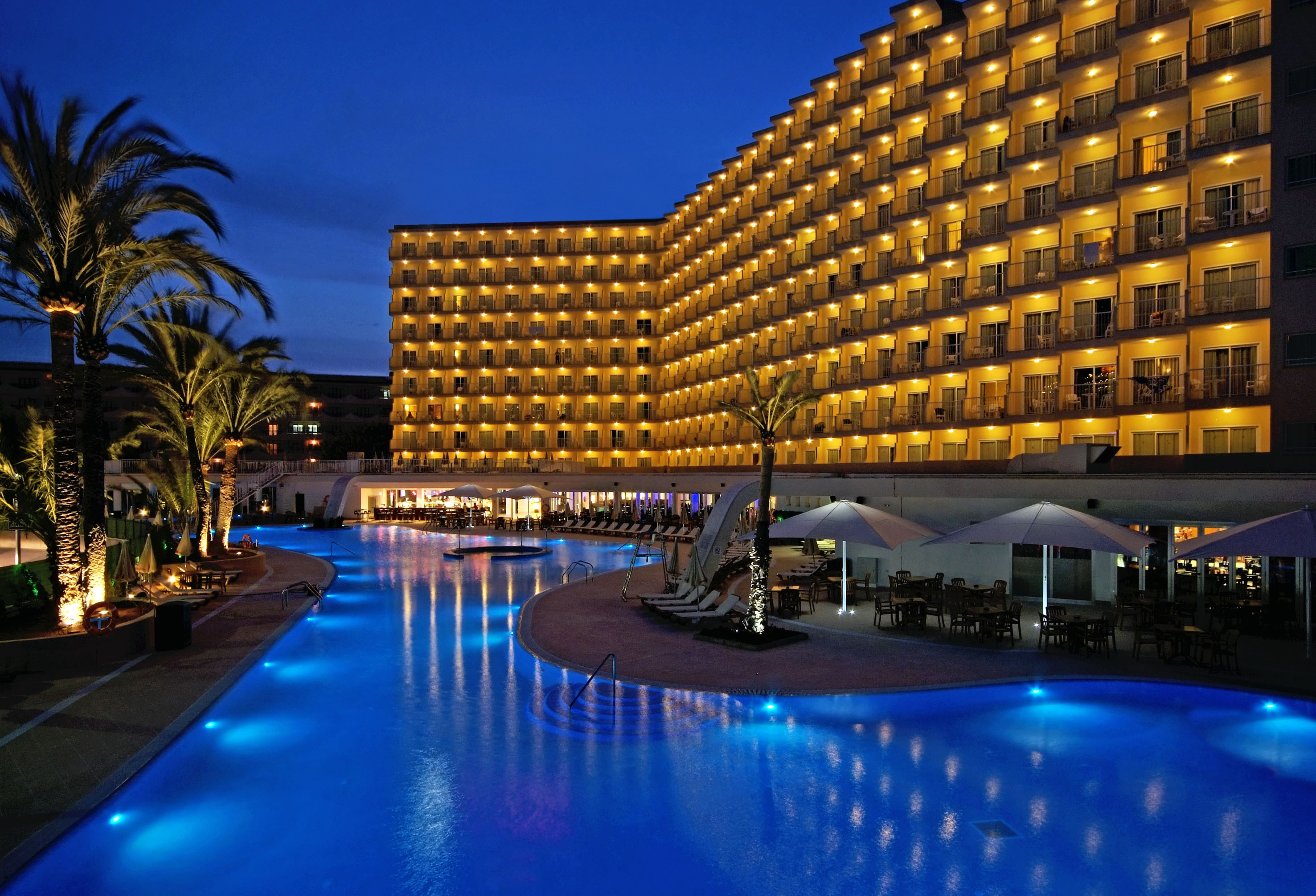 Hotel resort at night with illuminated pool, palm trees, and bright facade.