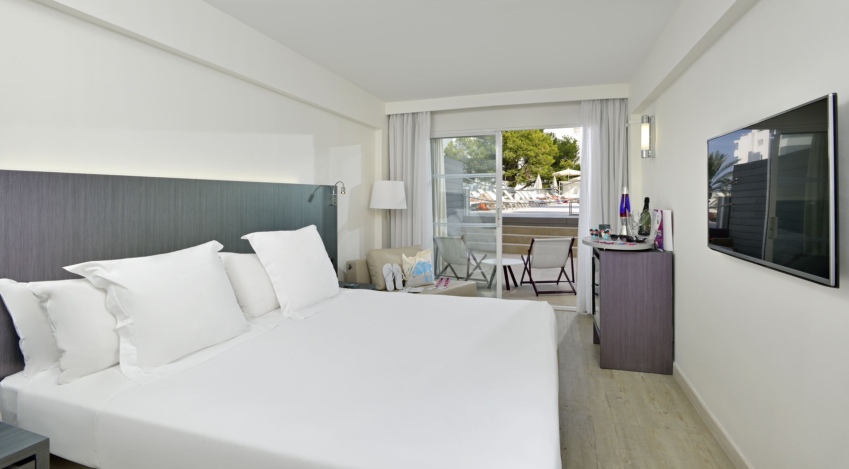Modern hotel room with white bed, balcony, pool view, and TV.