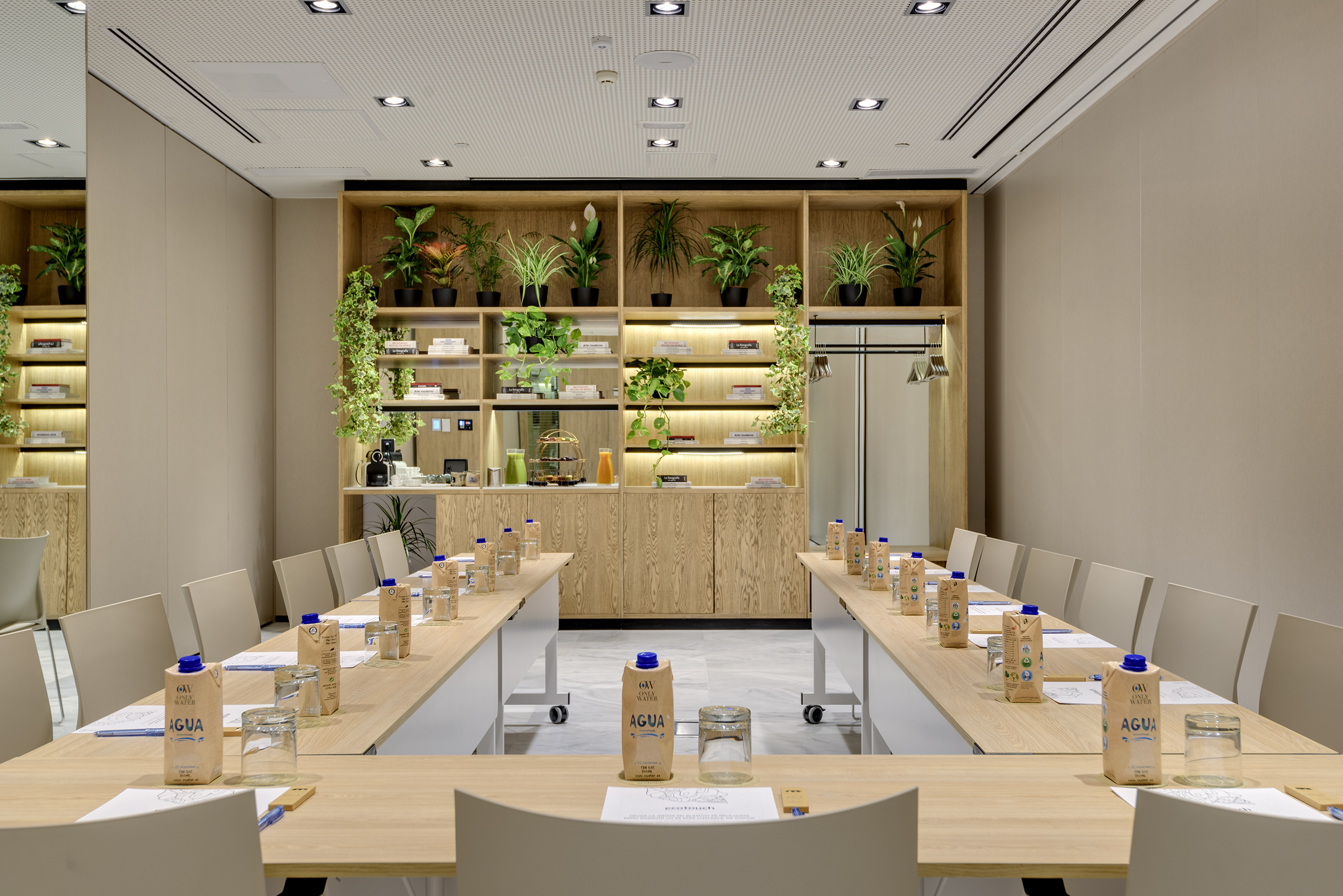 Modern meeting room with U-shaped tables, water, and wooden plant shelves.