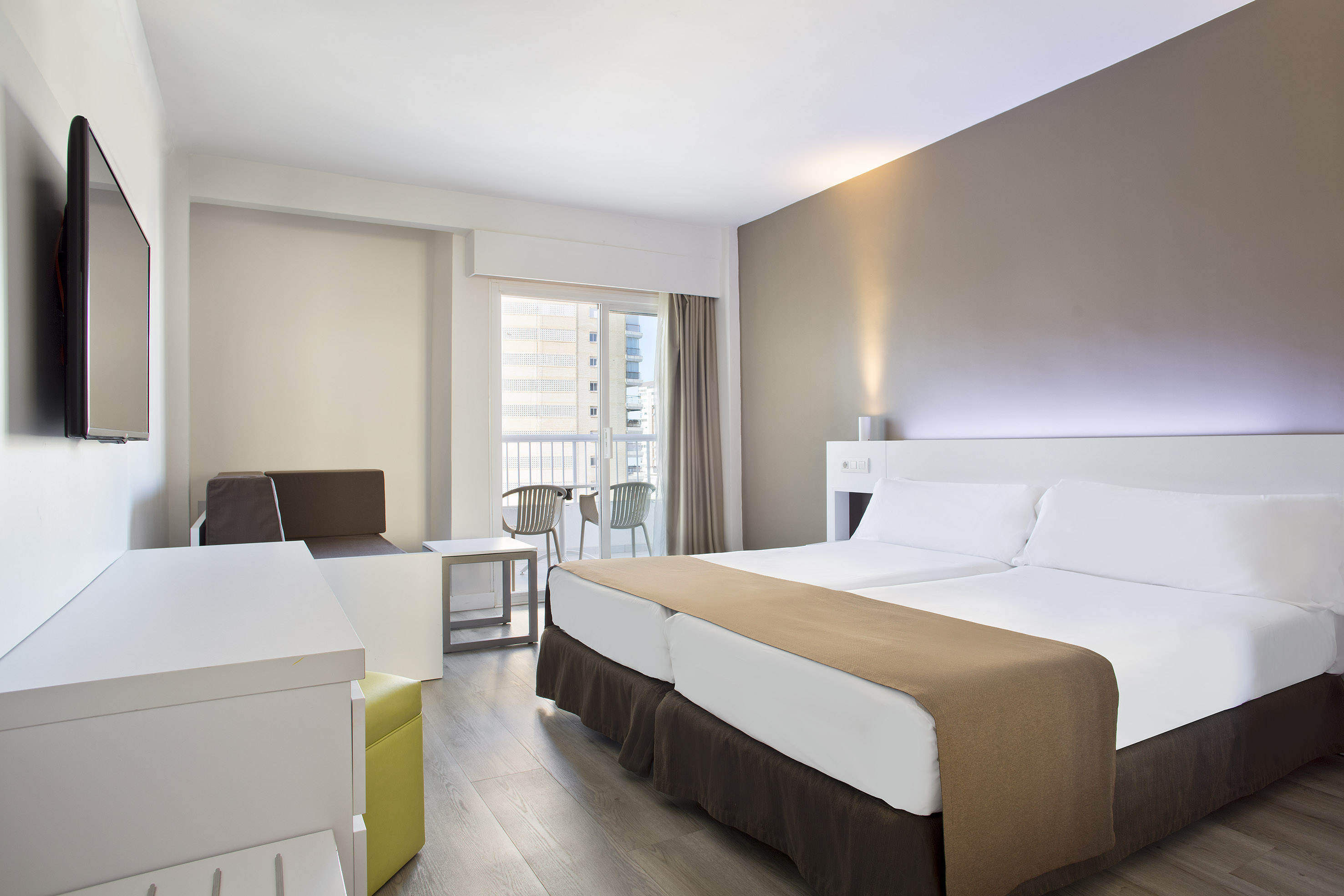 Modern hotel room with twin beds, a sofa, wall-mounted TV, and balcony.