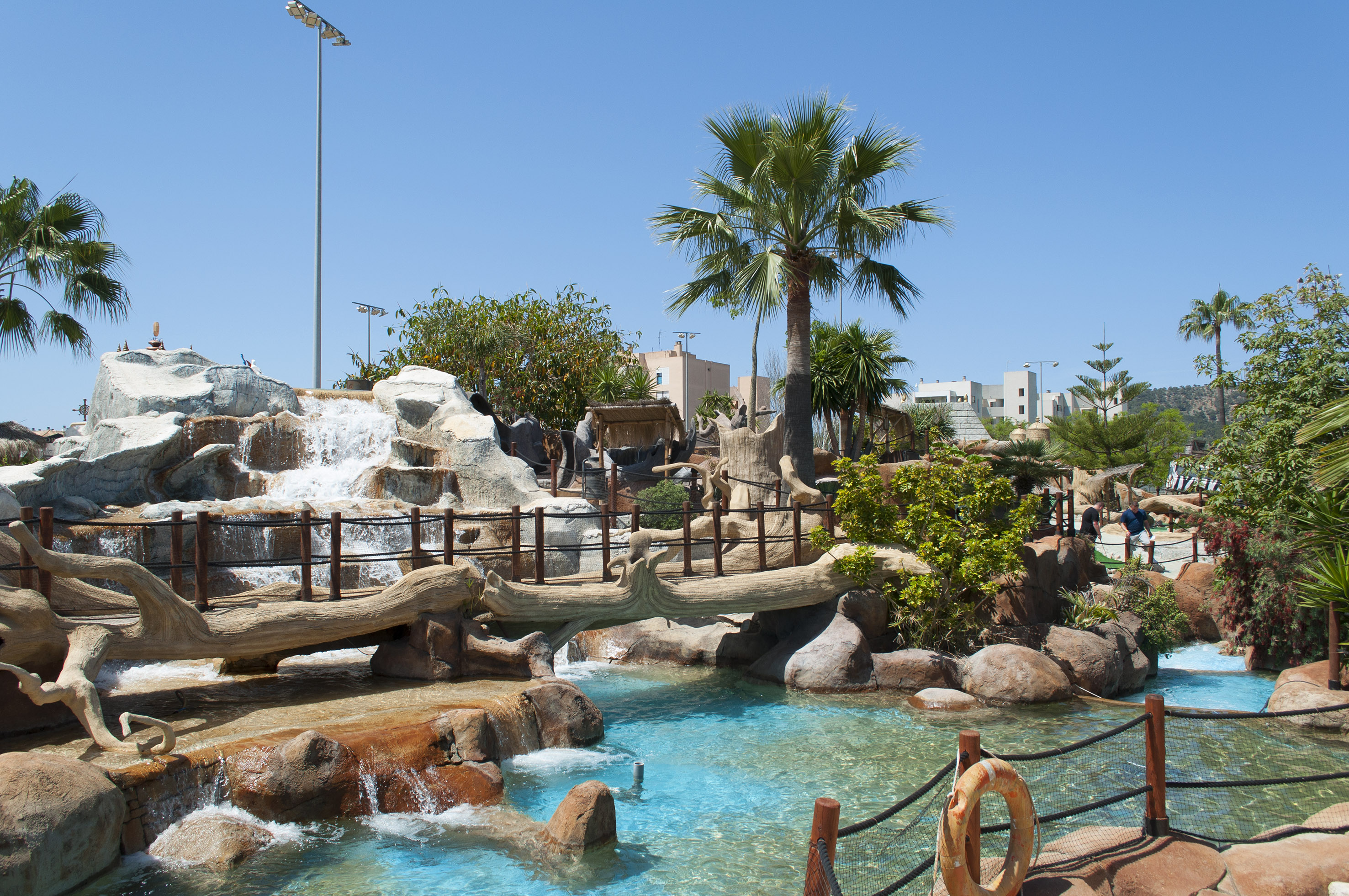 Water park waterfall, wooden bridge over blue water, lush palm trees.