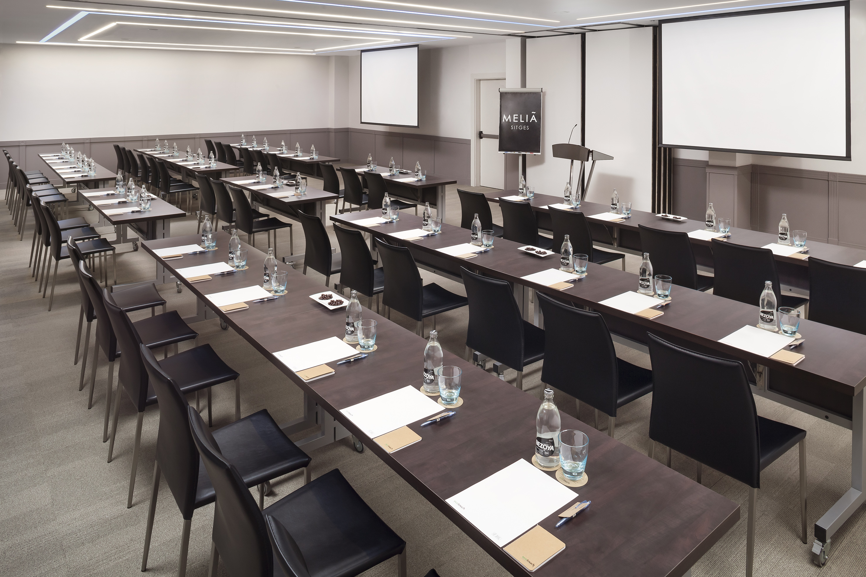 Meliã Sitges conference room: tables, chairs, dual screens, lectern setup.