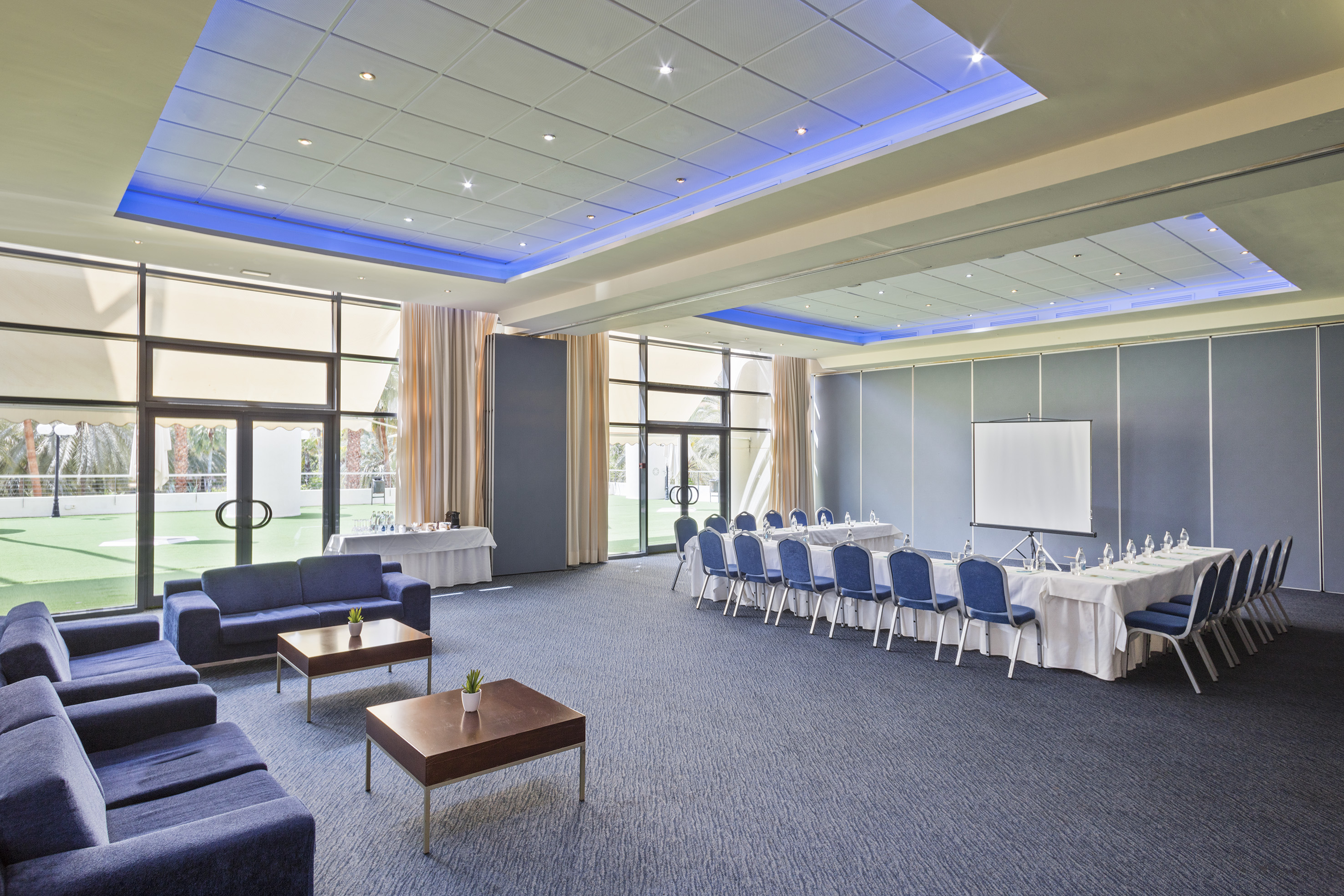 Modern meeting room with lounge, U-shaped tables, screen, and outdoor view.