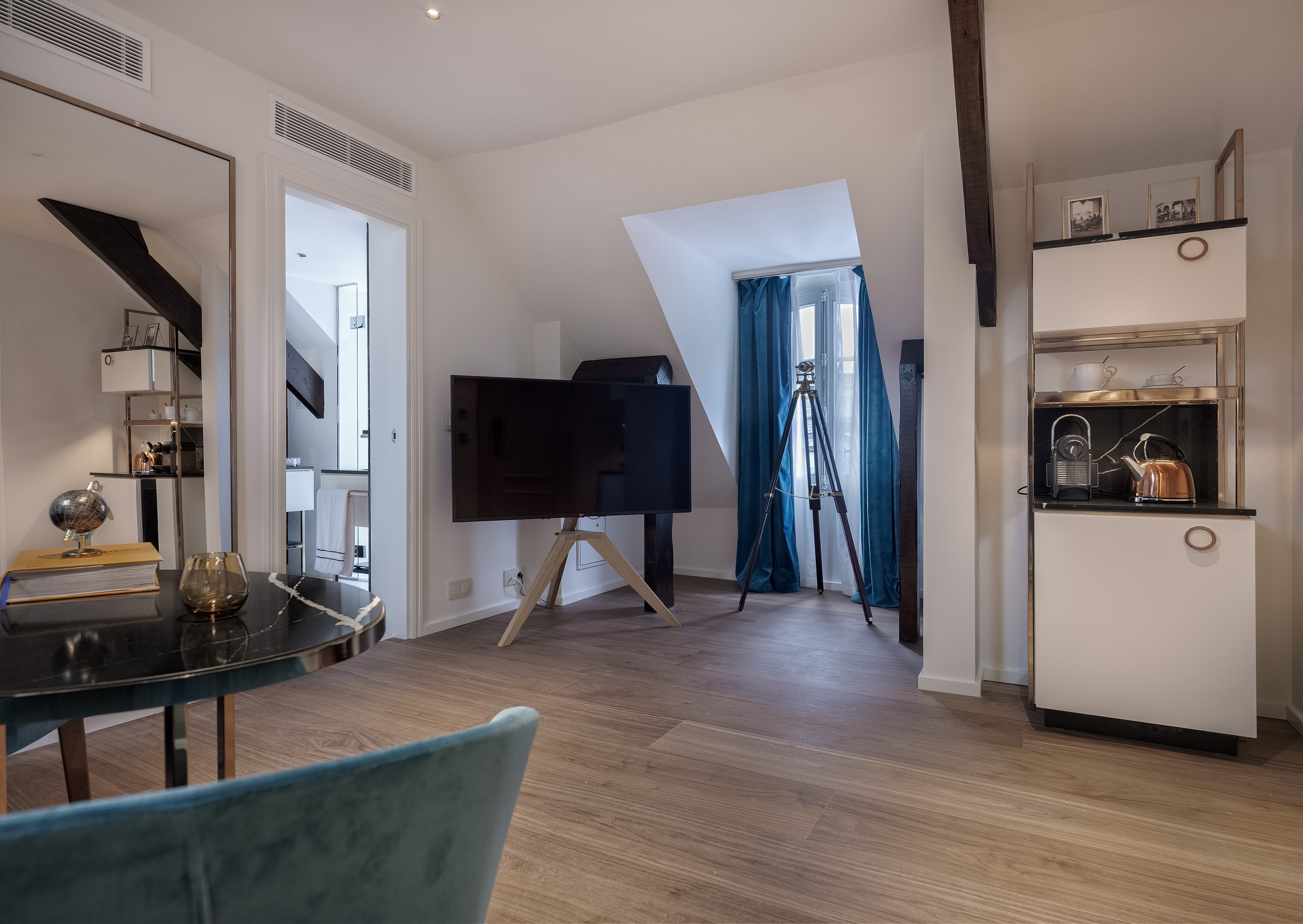 Modern apartment living area, TV, kitchenette, wood floor, blue curtains.