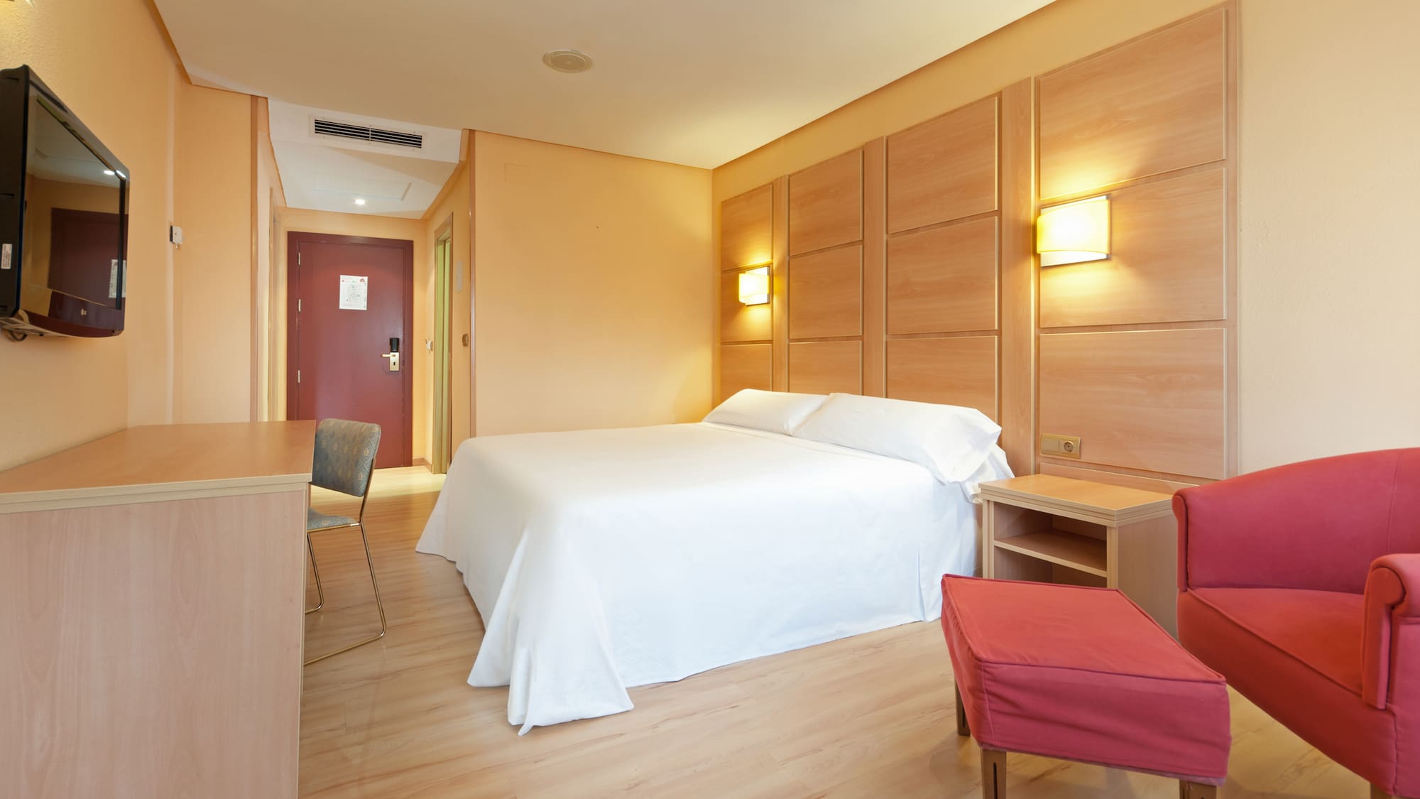Modern hotel room with white bed, wooden headboard, red armchair, and desk.