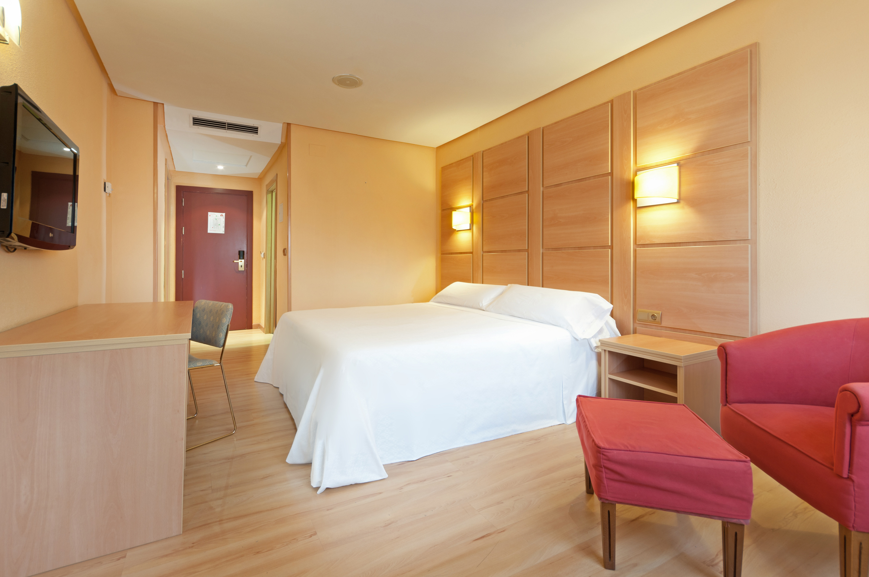 Modern hotel room with white bed, wooden headboard, red armchair, and desk.