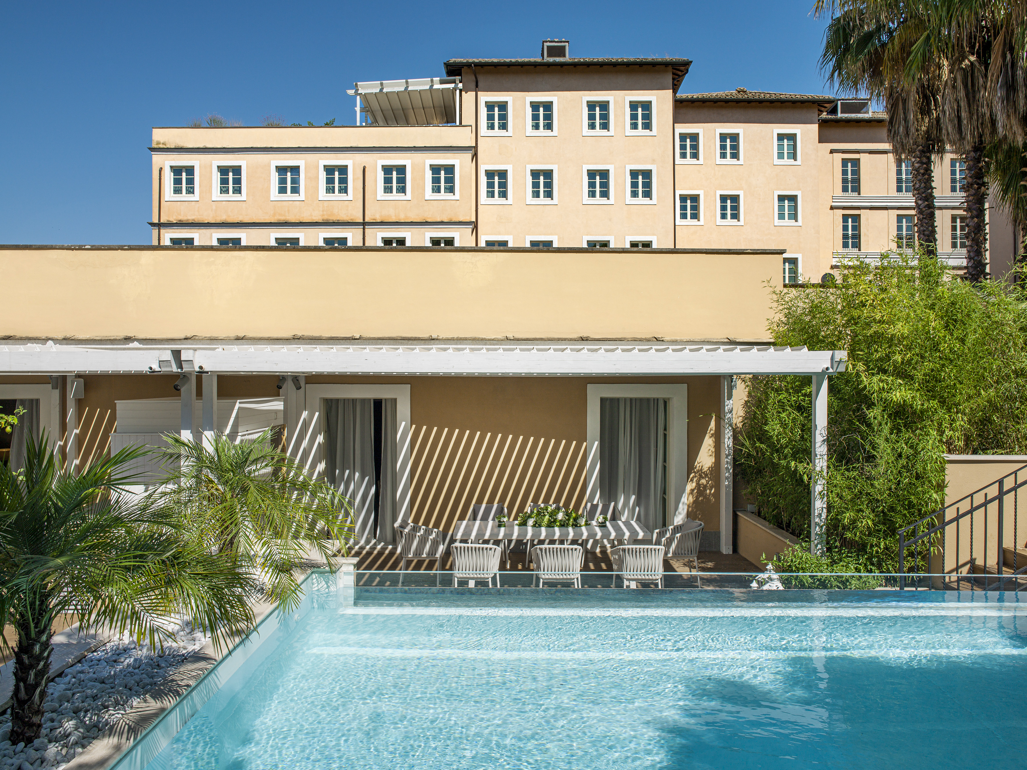 Sunny hotel pool and patio with outdoor dining and palm trees.