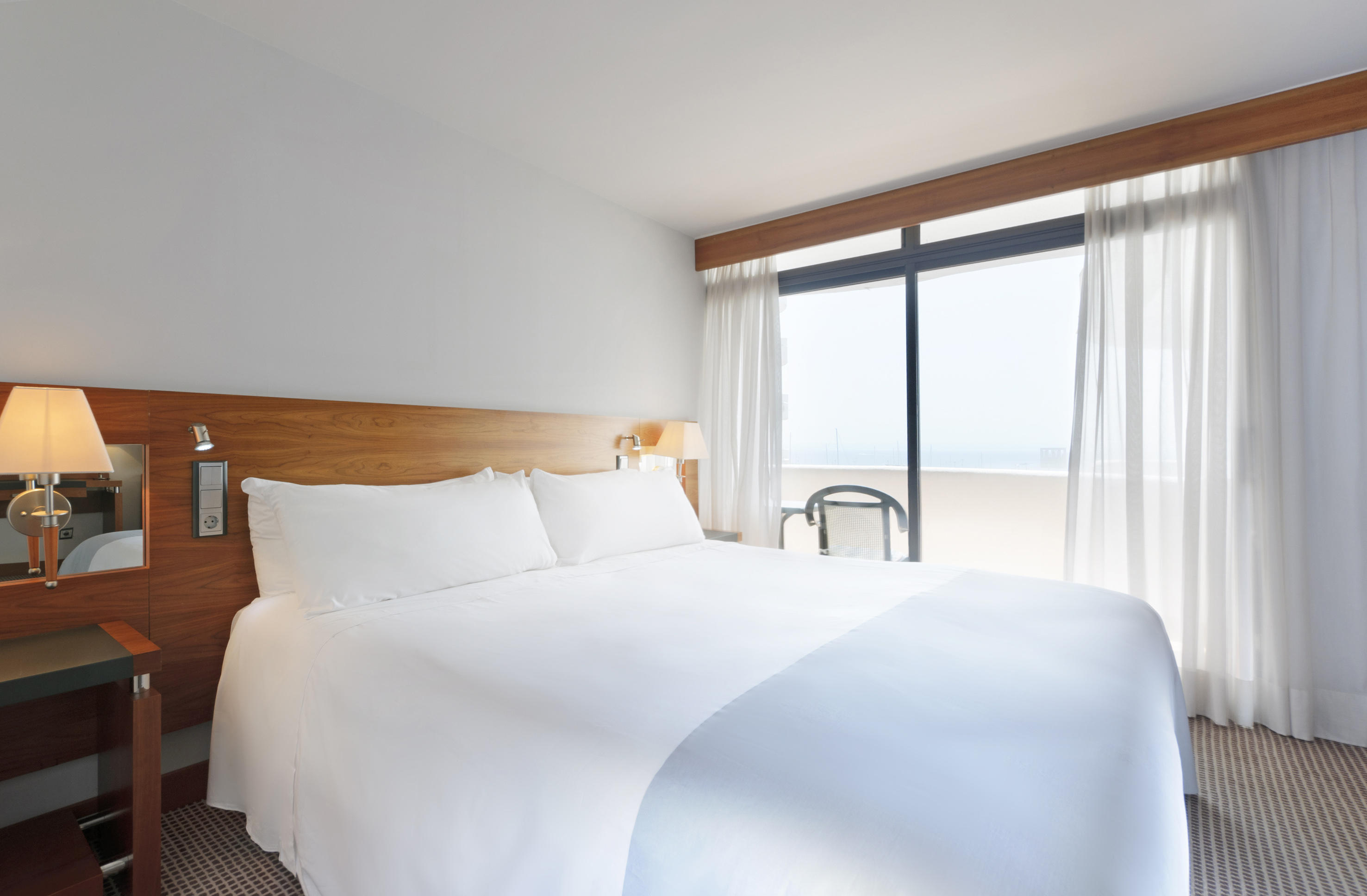 White hotel bedroom with king bed, wooden headboard, and balcony window.