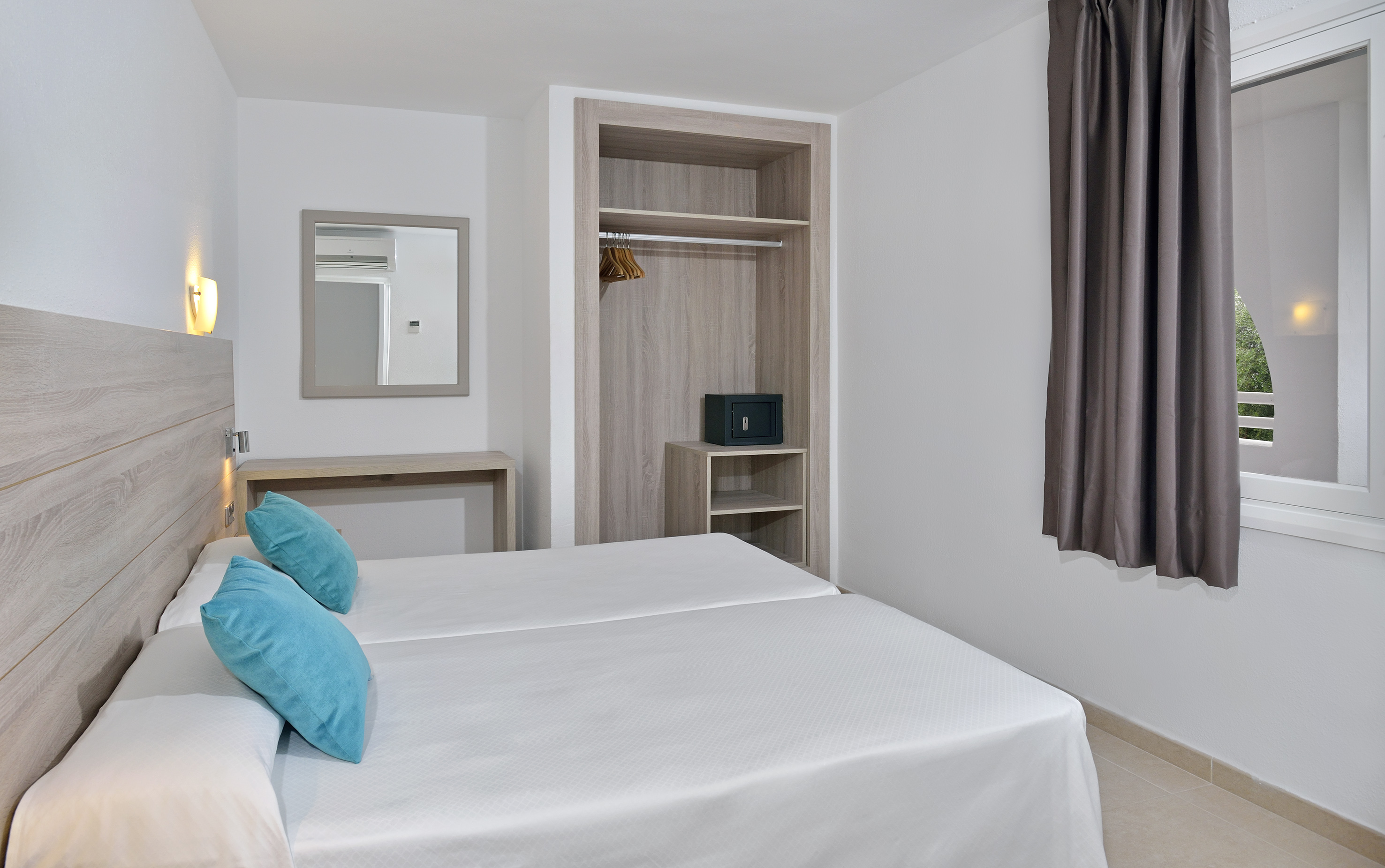 Twin beds in a modern hotel room with open wardrobe, safe, and mirror.