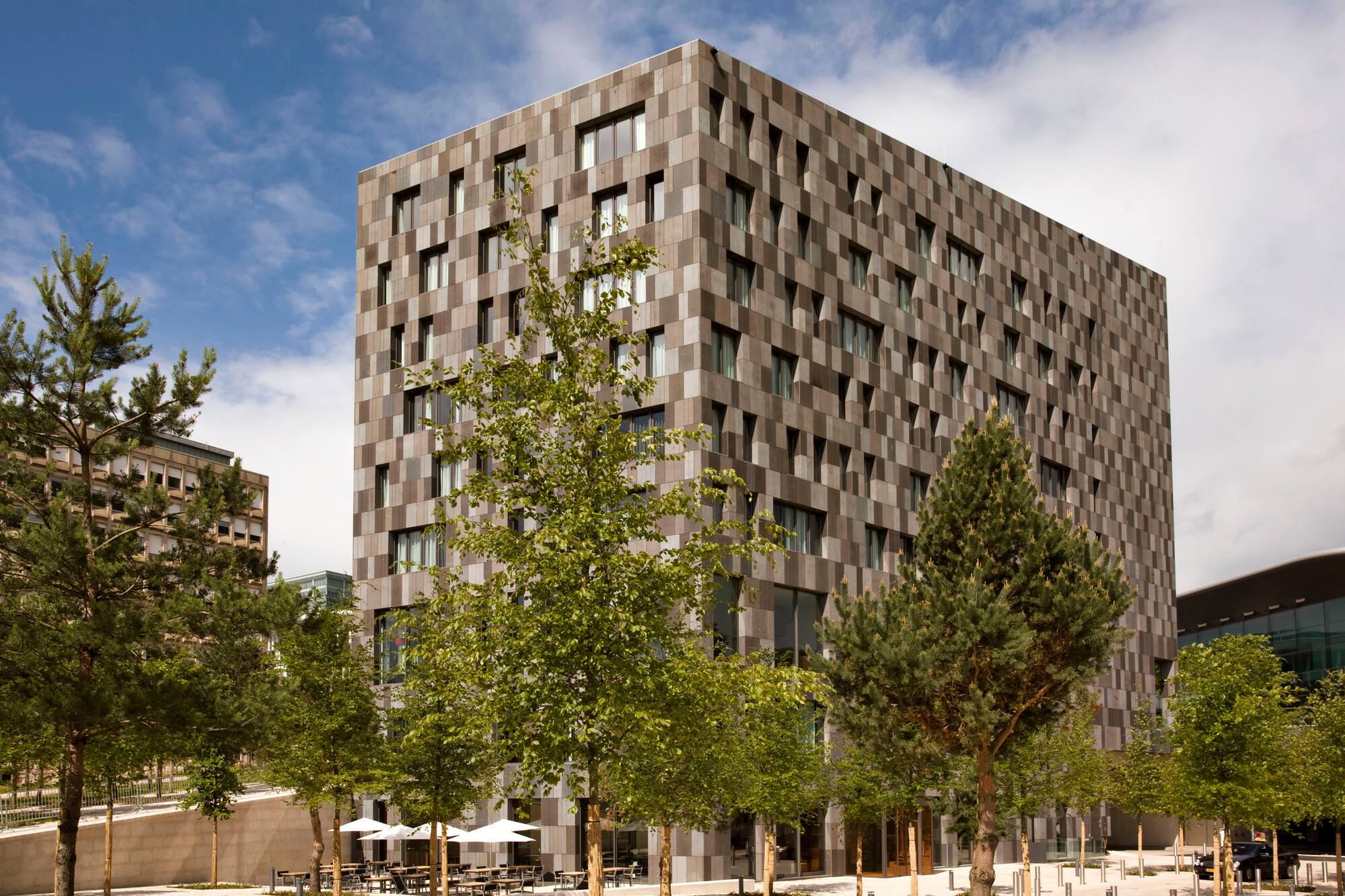 Modern building with gray checkered facade, green trees, and outdoor cafe area.