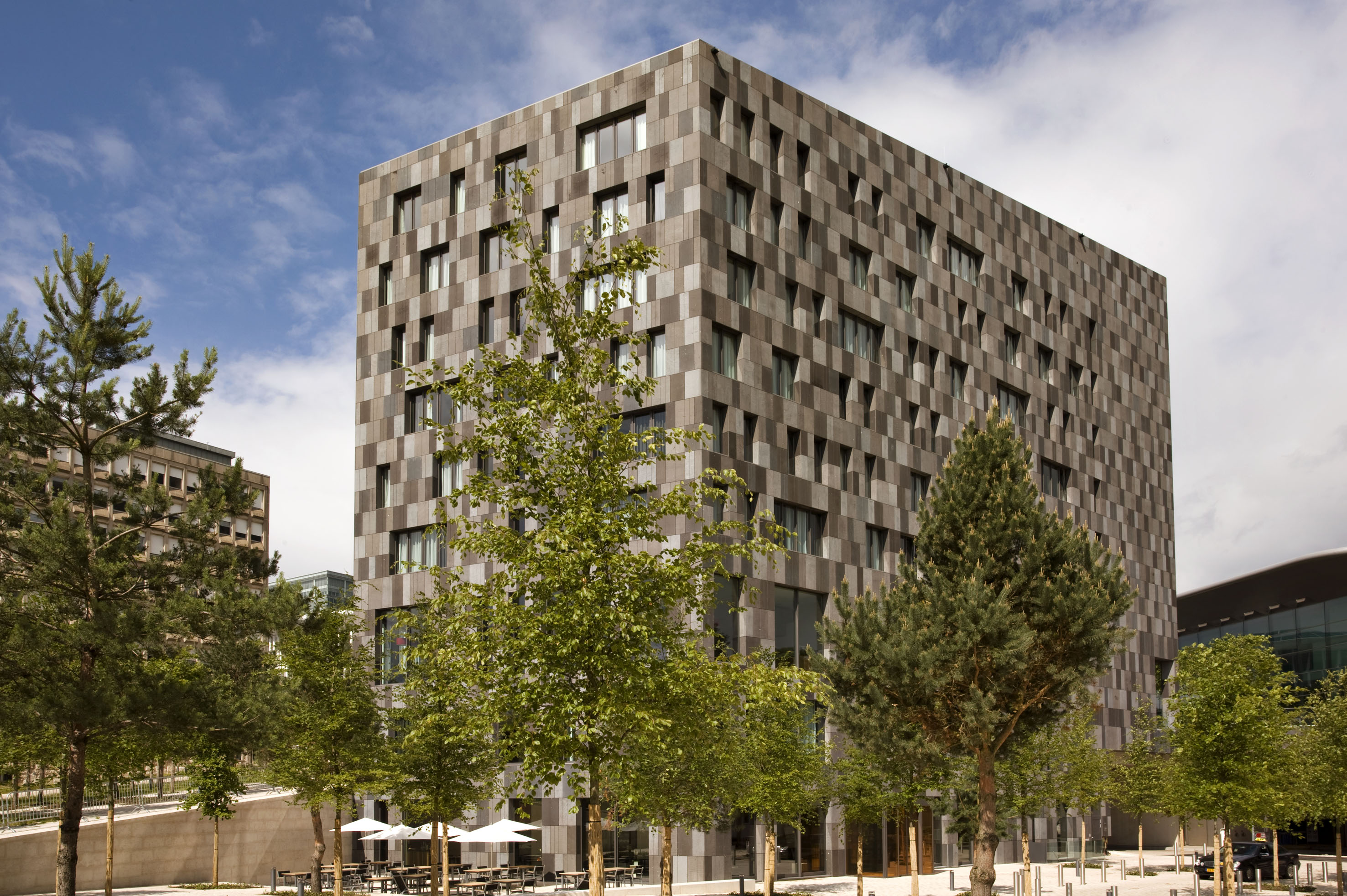 Modern building with gray checkered facade, green trees, and outdoor cafe area.