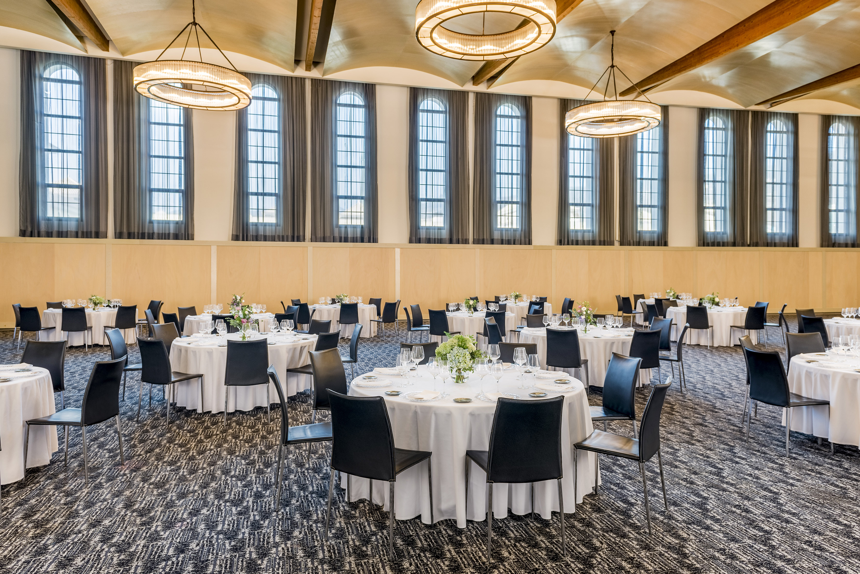Event space set with round tables, black chairs, arched windows, and chandeliers.