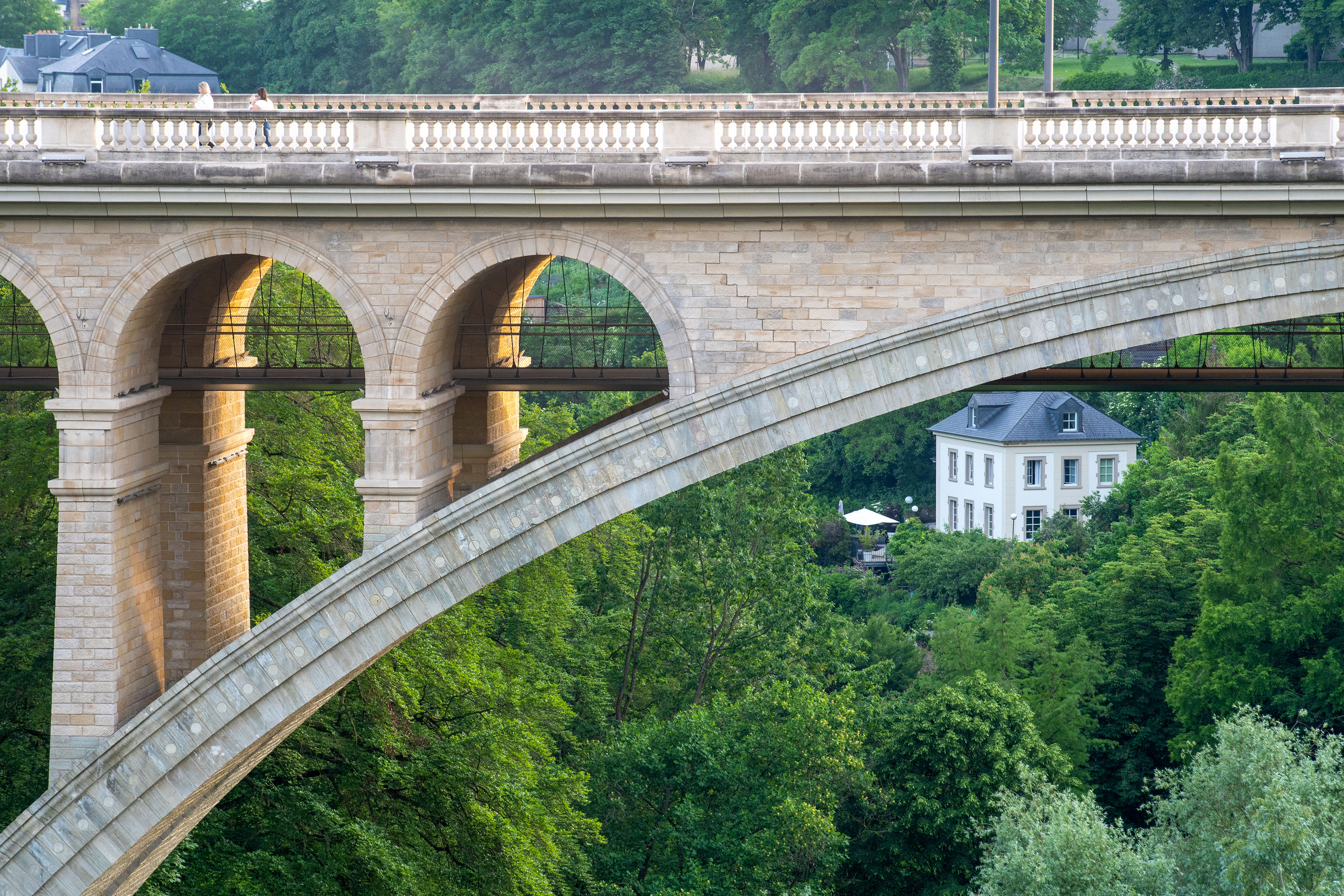 Stone viaduct bridge arches over green forest; white house and people visible.