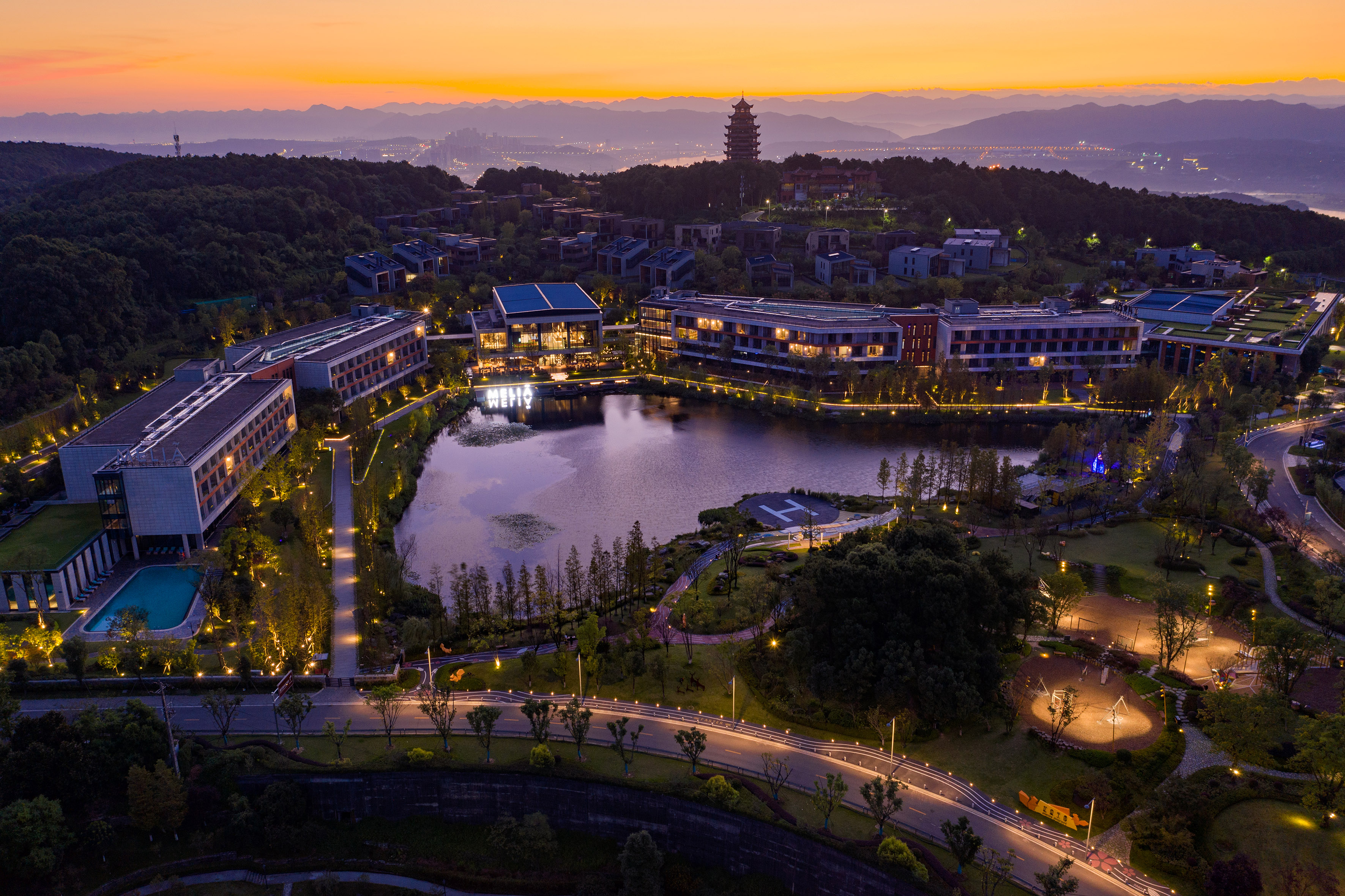 Illuminated modern resort complex, lake, and pagoda under sunset sky.