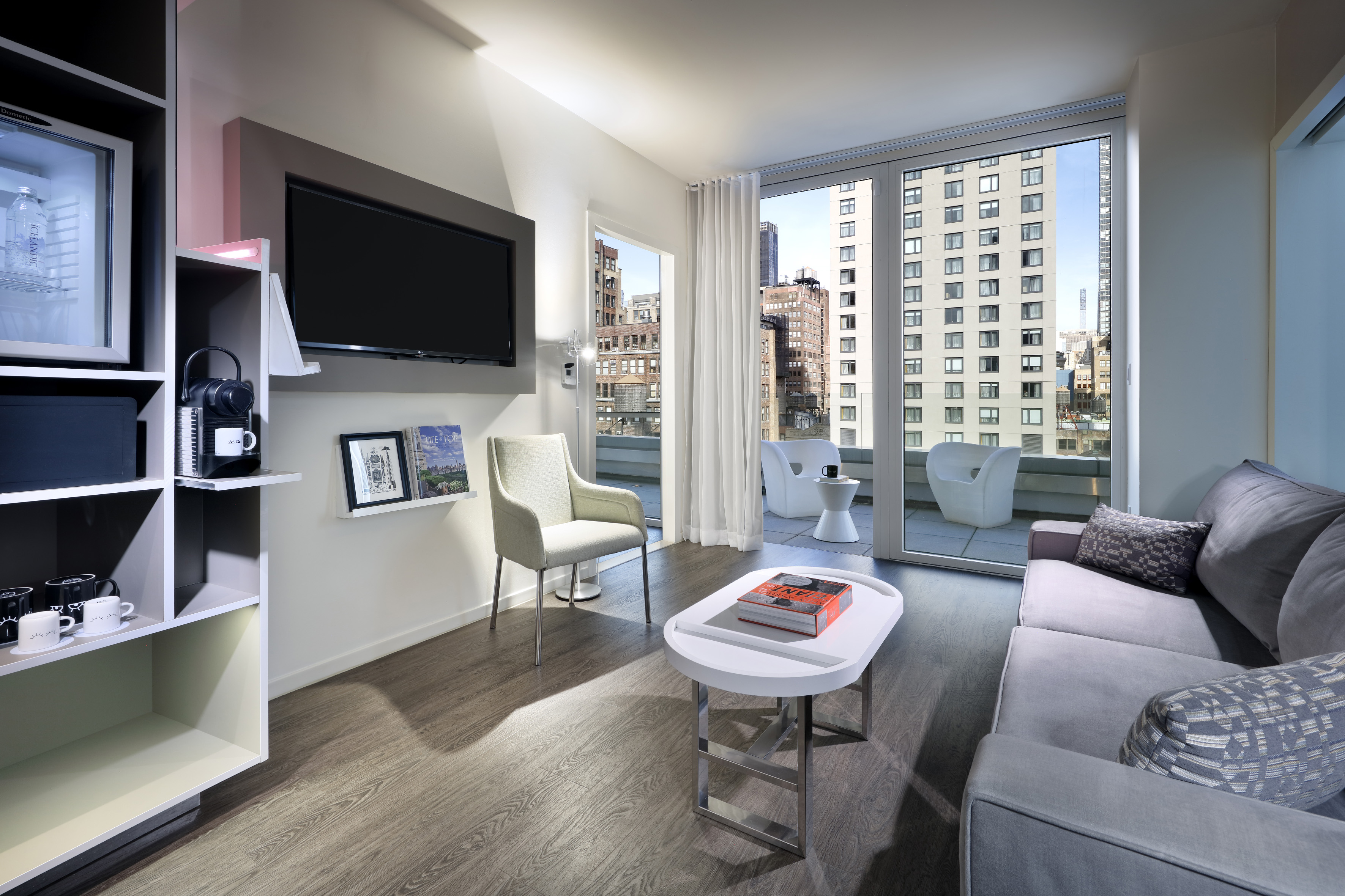 Modern hotel living room with balcony, city view, grey sofa, and mounted TV.
