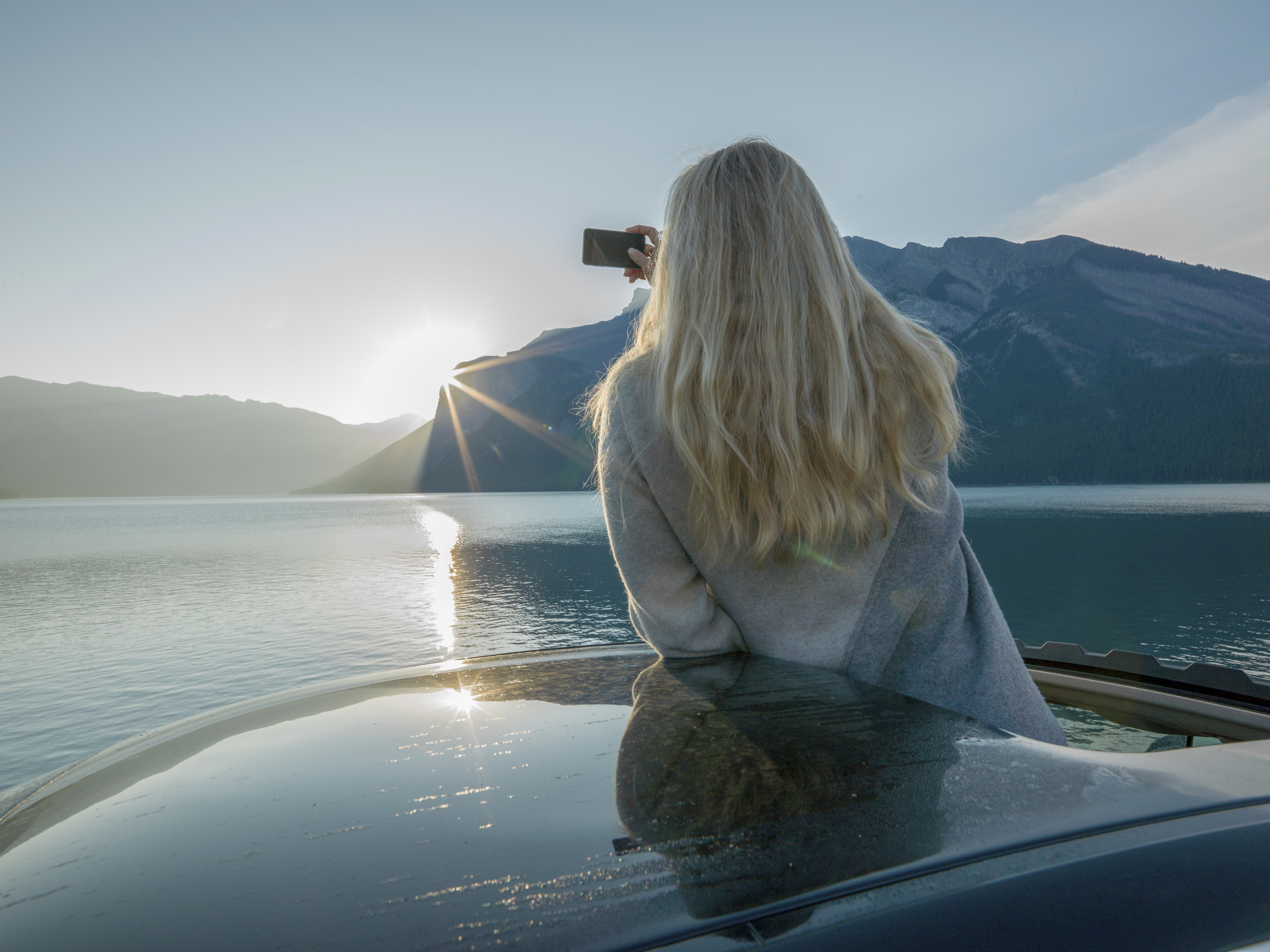 a woman sitting on a car taking a picture of a mountain range