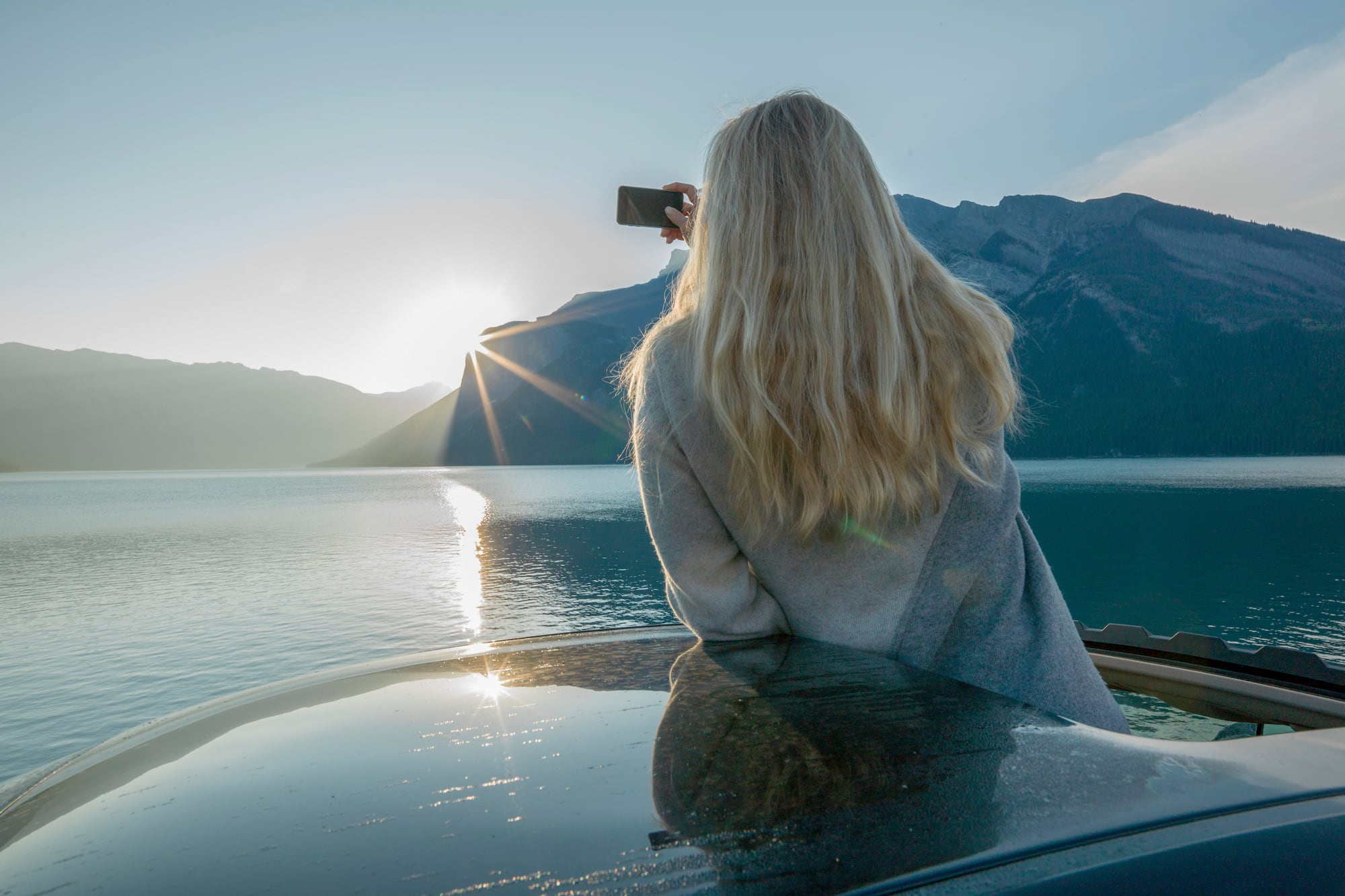 a woman sitting on a car taking a picture of a mountain range