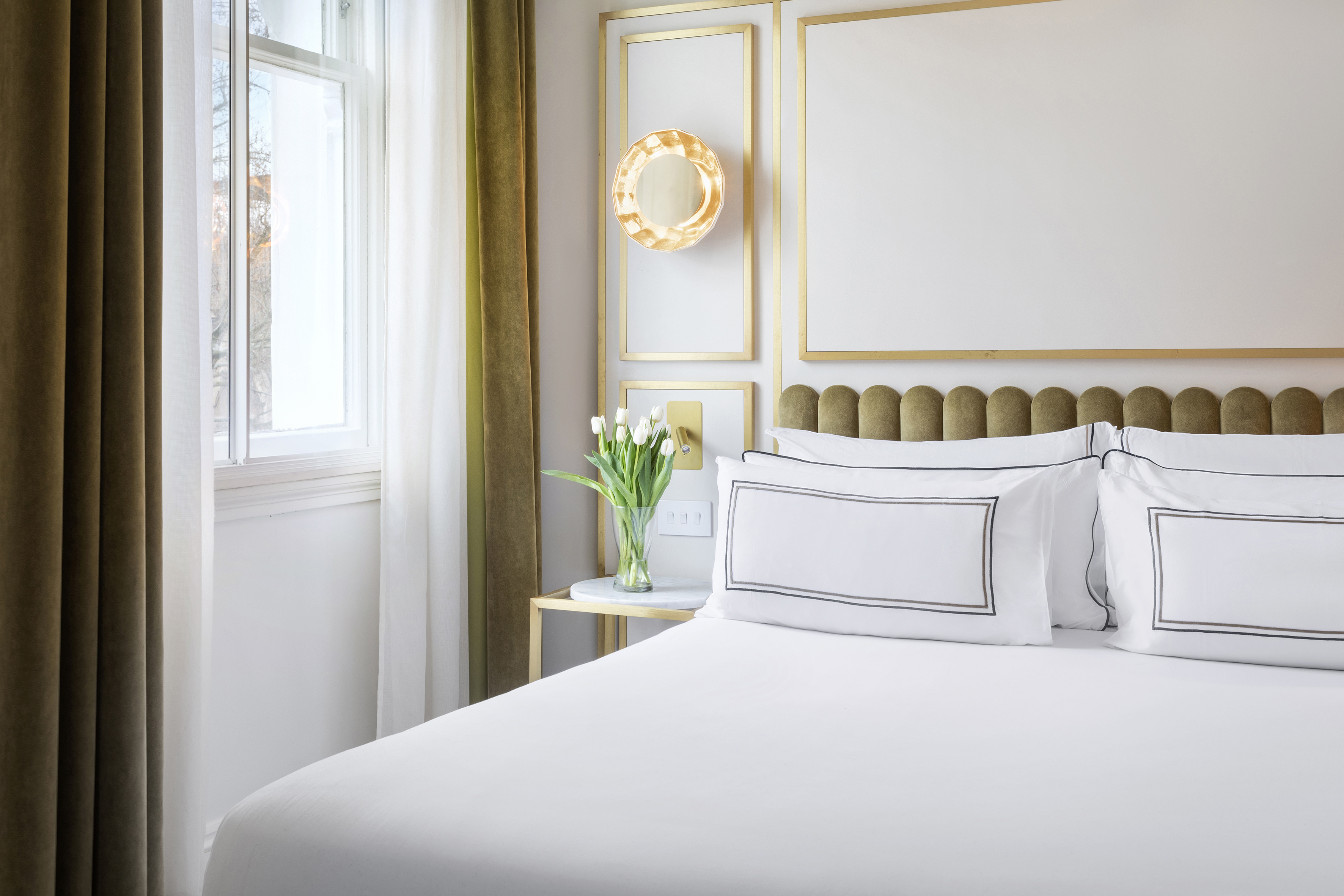 Modern bedroom with white bed, olive headboard, gold decor, and tulips.
