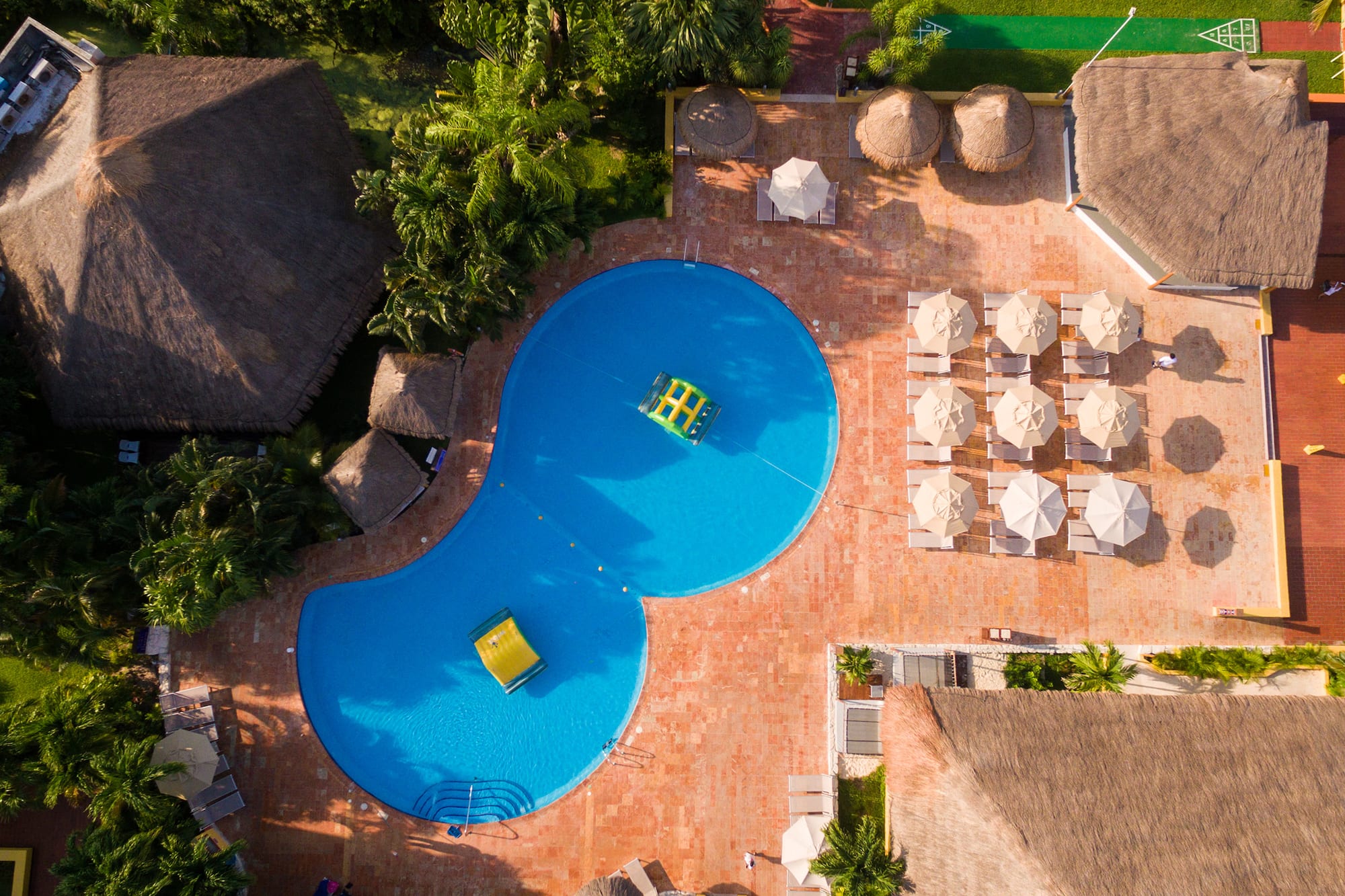Resort tropical con piscina azul, palapas, tumbonas y palmeras desde el aire.