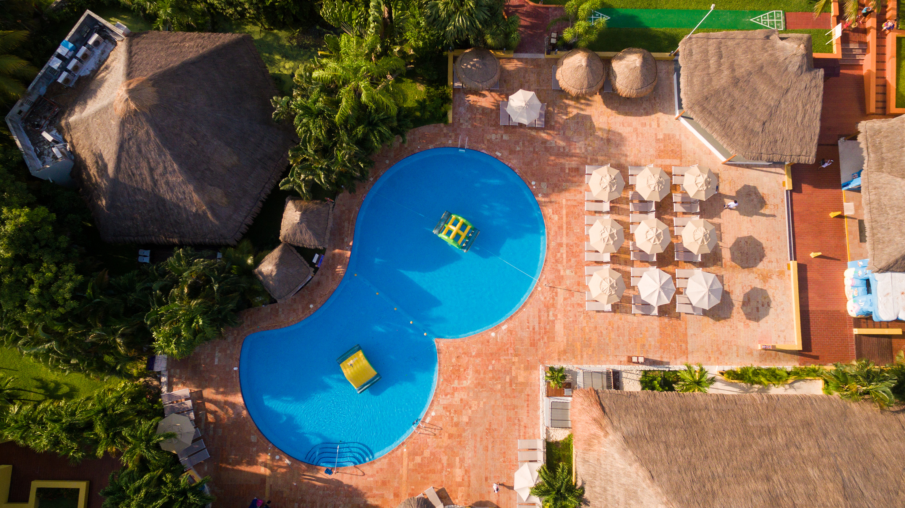 Resort tropical con piscina azul, palapas, tumbonas y palmeras desde el aire.