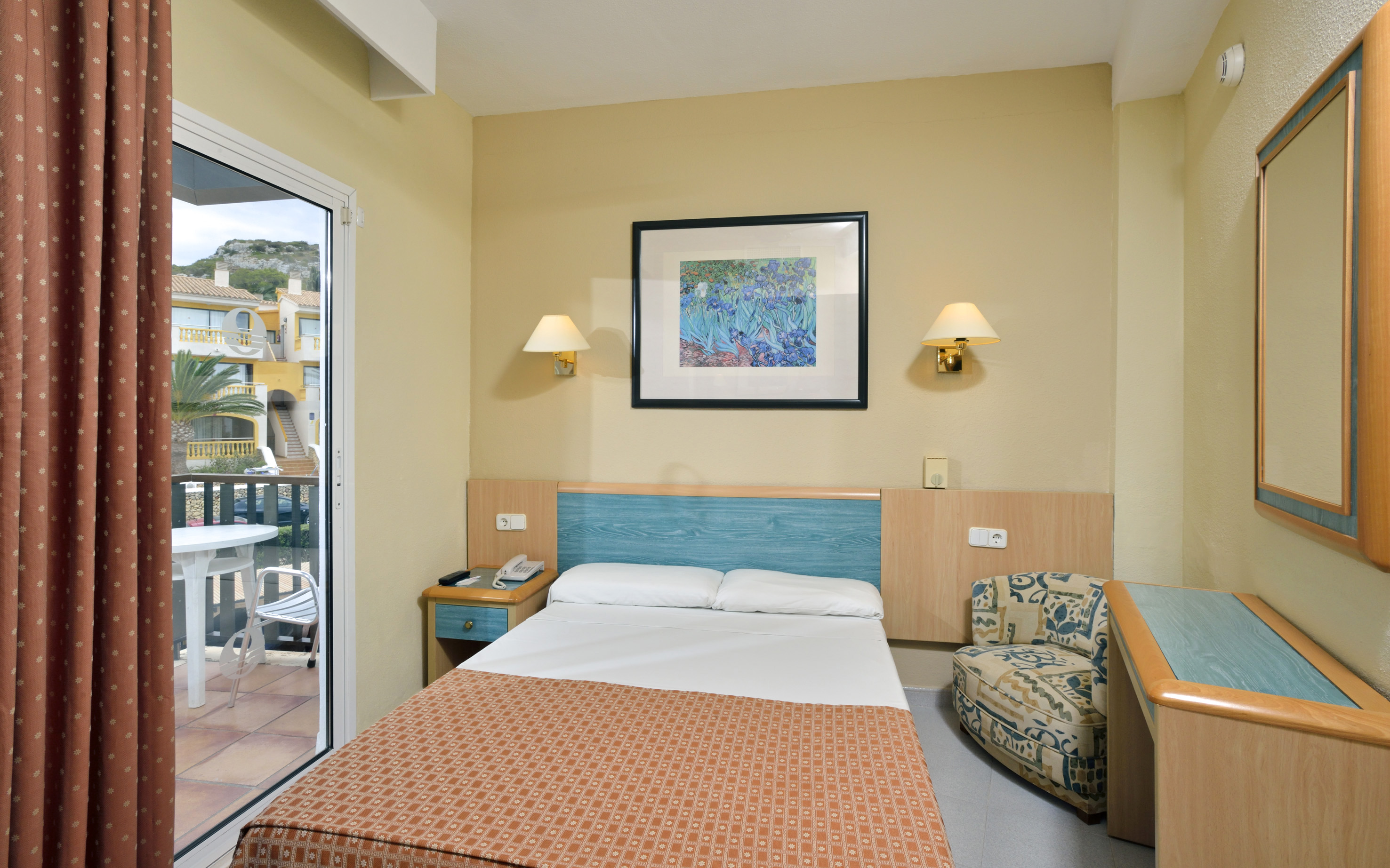 Hotel single room with bed, armchair, desk, balcony, and outdoor view.