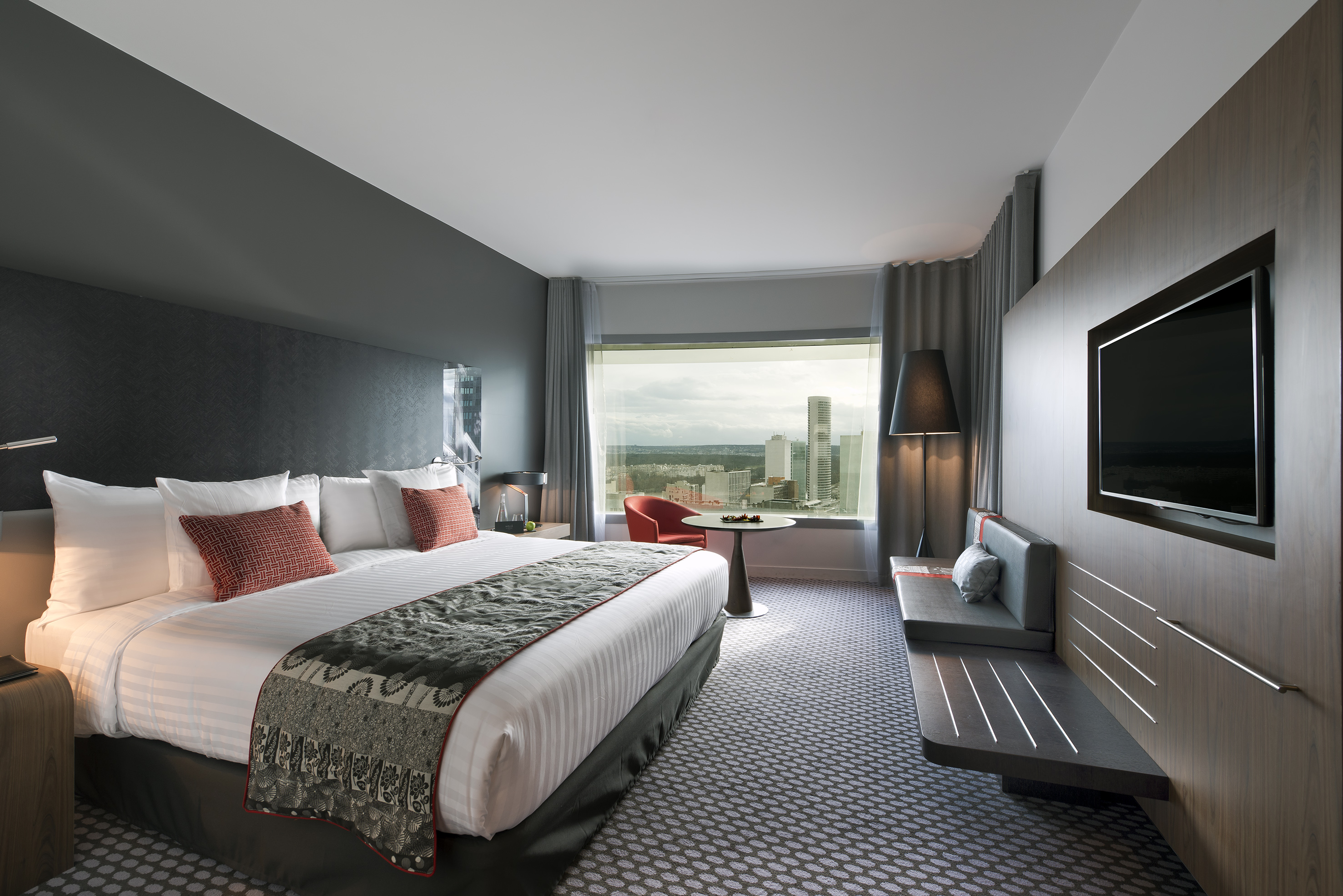 Modern hotel room with city view, large bed, and wall-mounted TV.