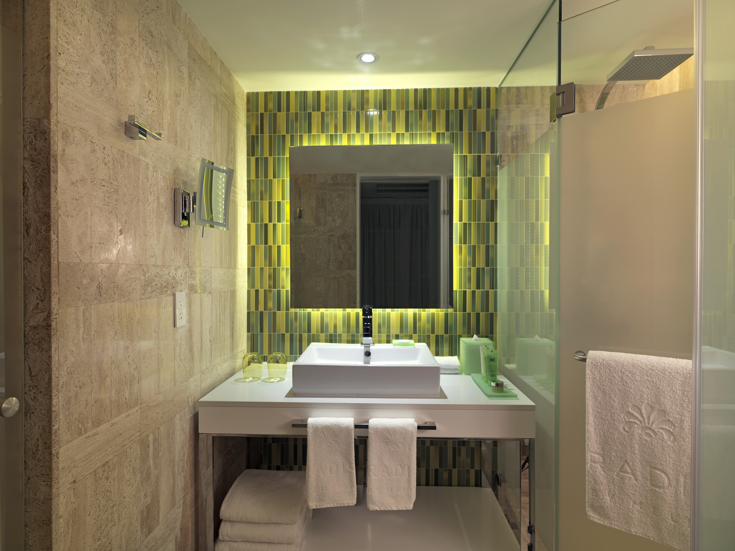 Contemporary hotel bathroom with square vanity, backlit mirror, mosaic tiles, shower.