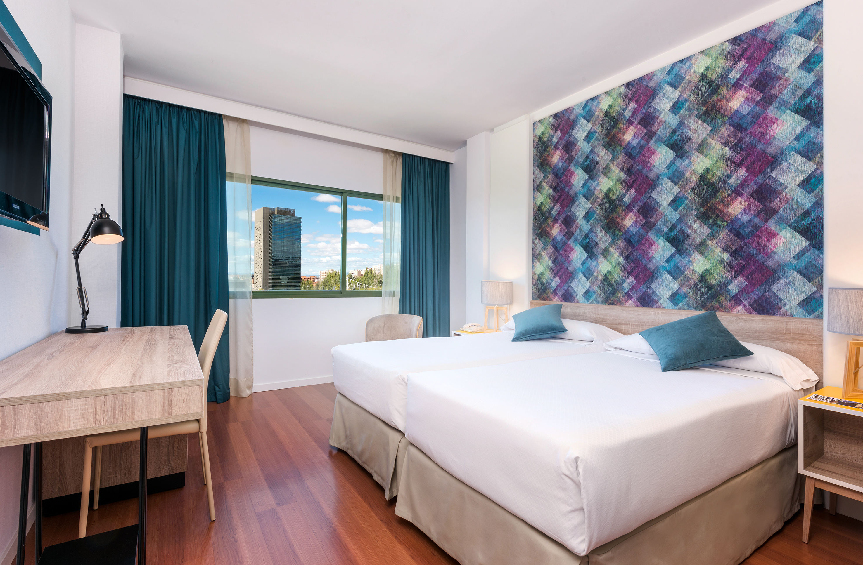 Modern hotel room with twin beds, colorful accent wall, desk, and city view.