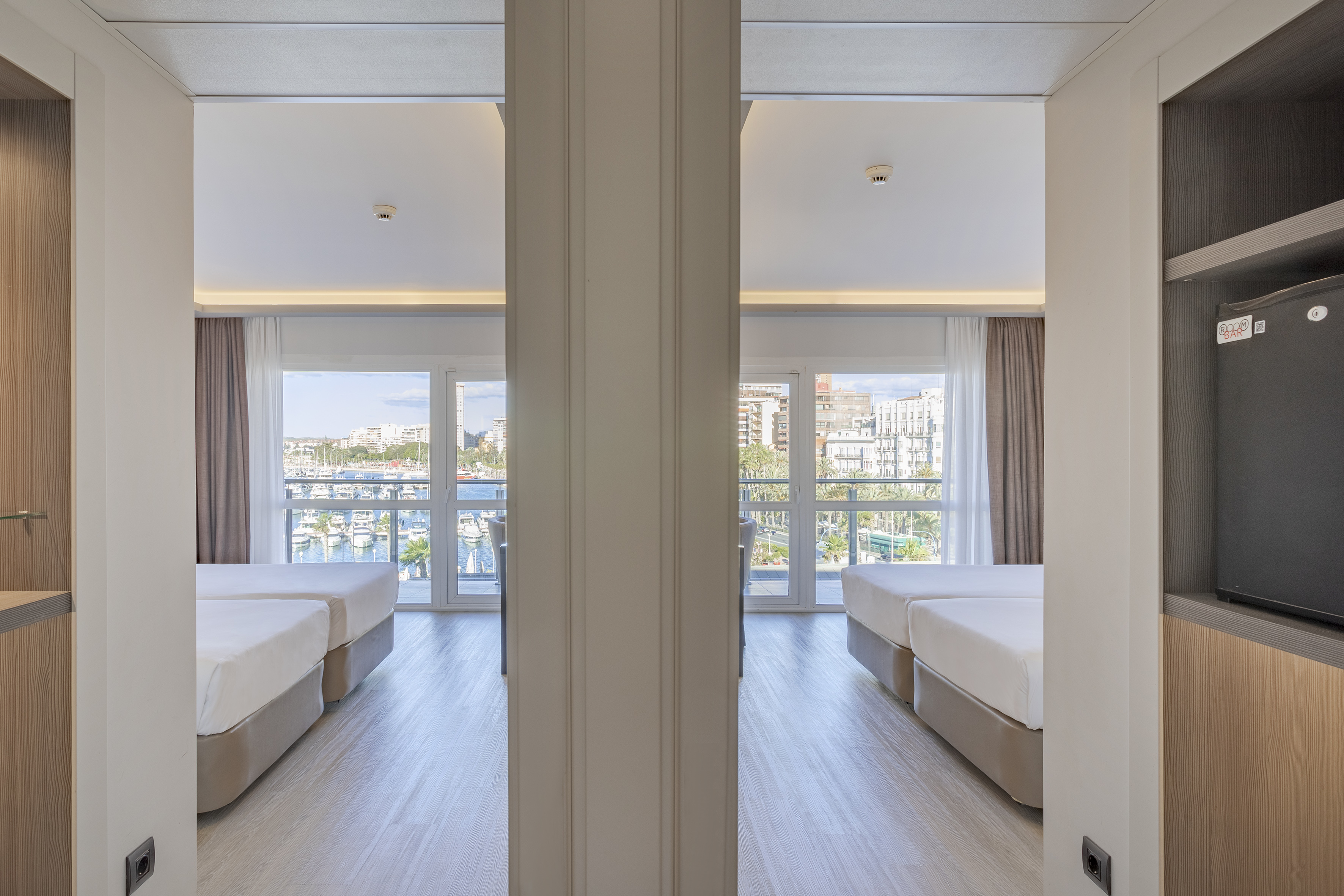 Bright hotel twin room with two beds, large windows, and a marina view.