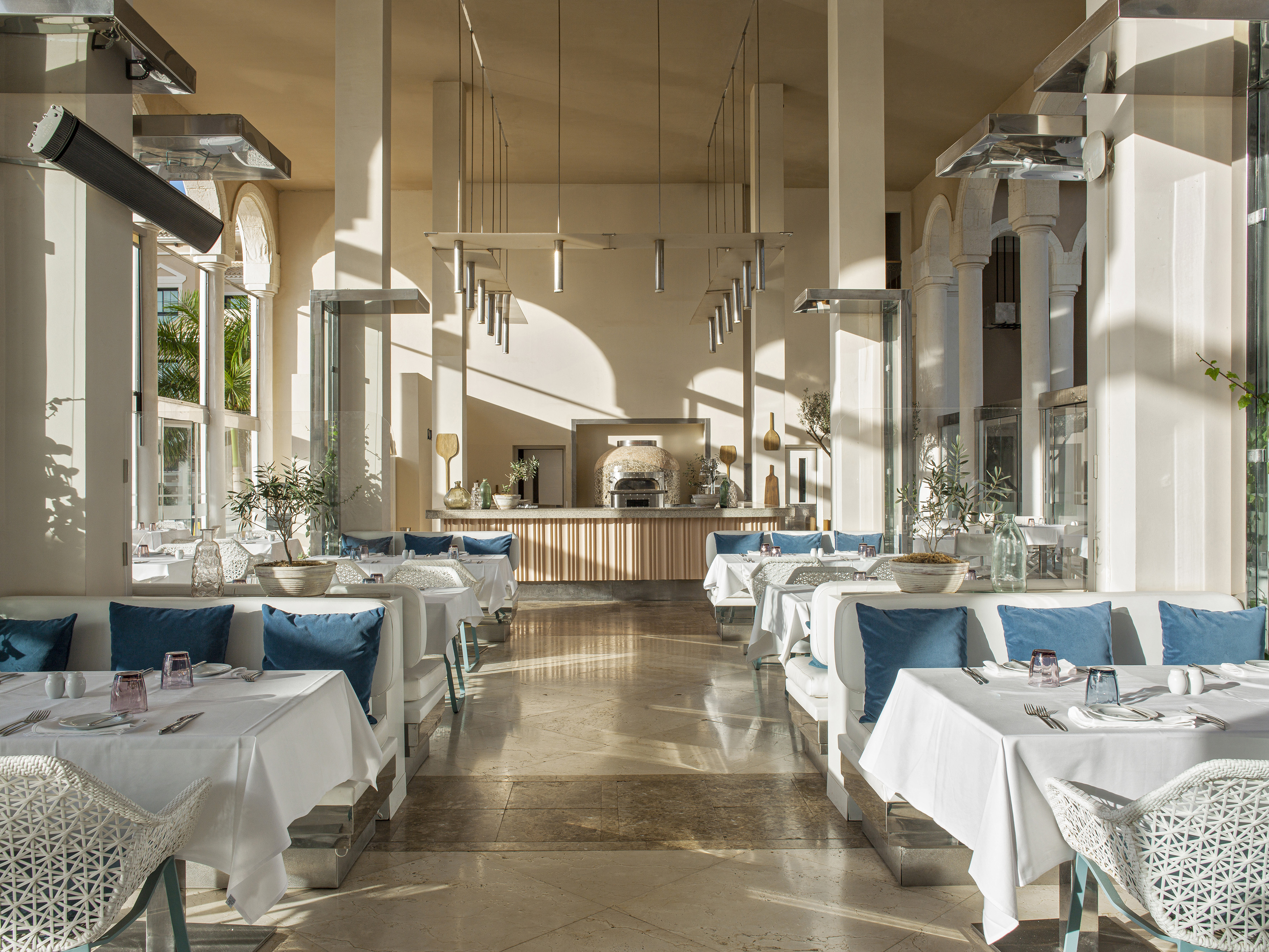 Sunny modern restaurant with white tables, blue cushions, and a pizza oven.