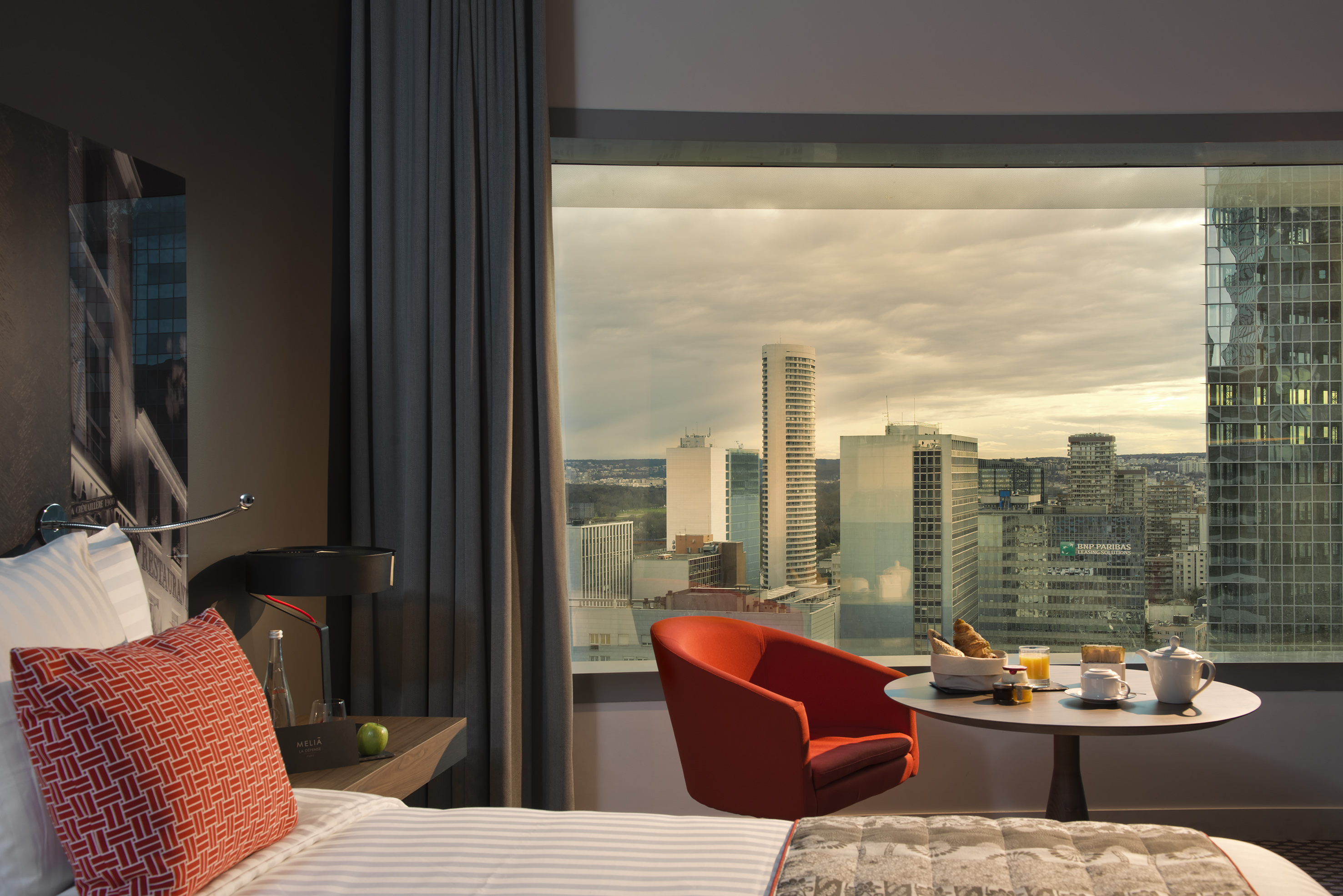 Modern hotel room with city skyline view, breakfast table, and orange armchair.